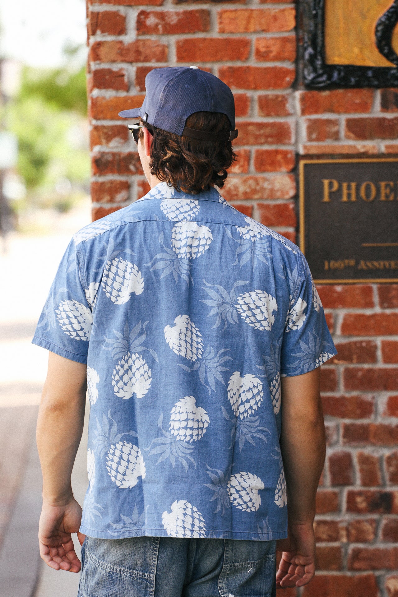 Man wearing Duke Kahanamoku indigo cotton Hawaiian shirt with white pineapple print, standing by brick wall