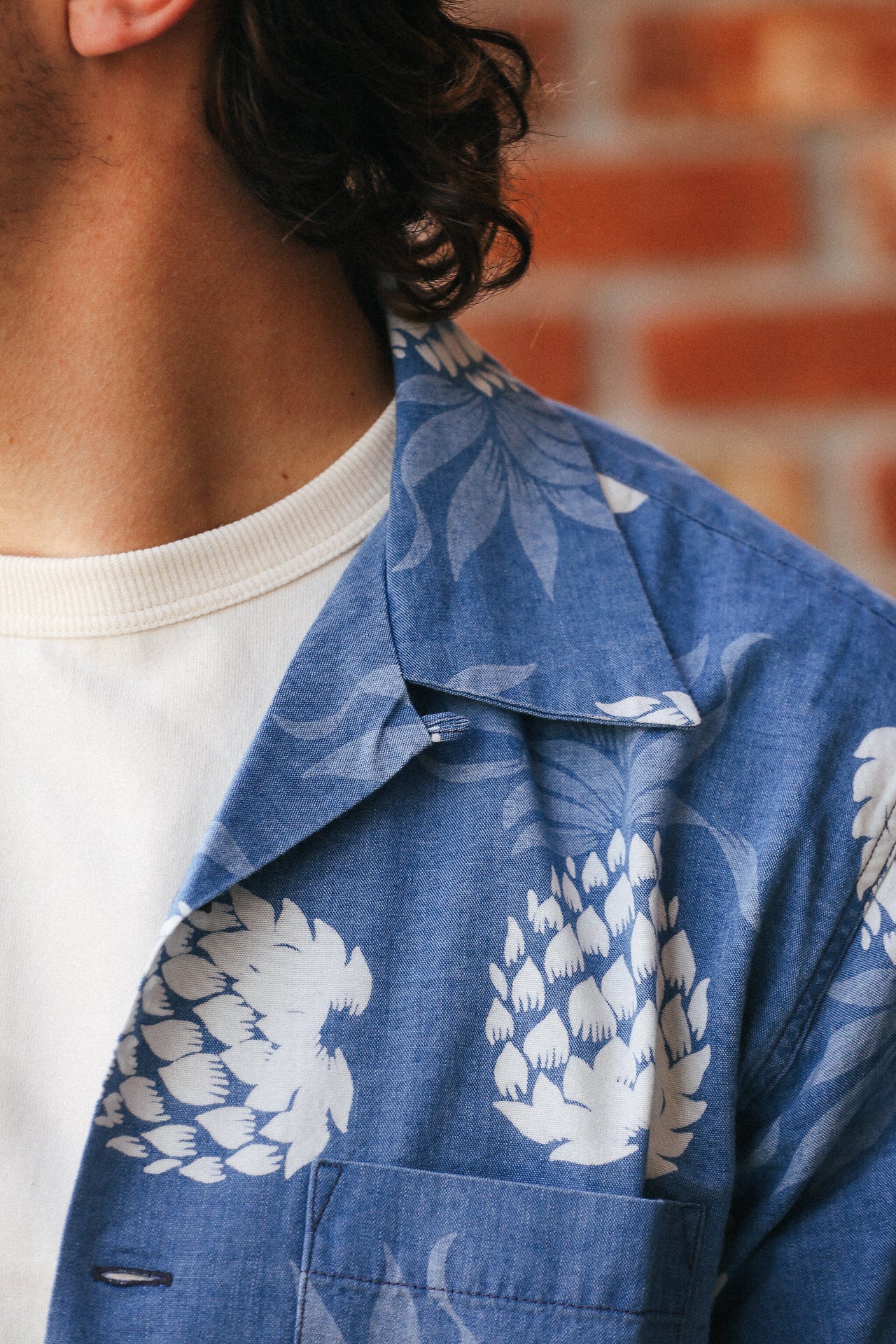 Close-up of a person wearing a blue Duke Kahanamoku indigo cotton Hawaiian shirt with white pineapple prints over a white ribbed crewneck shirt