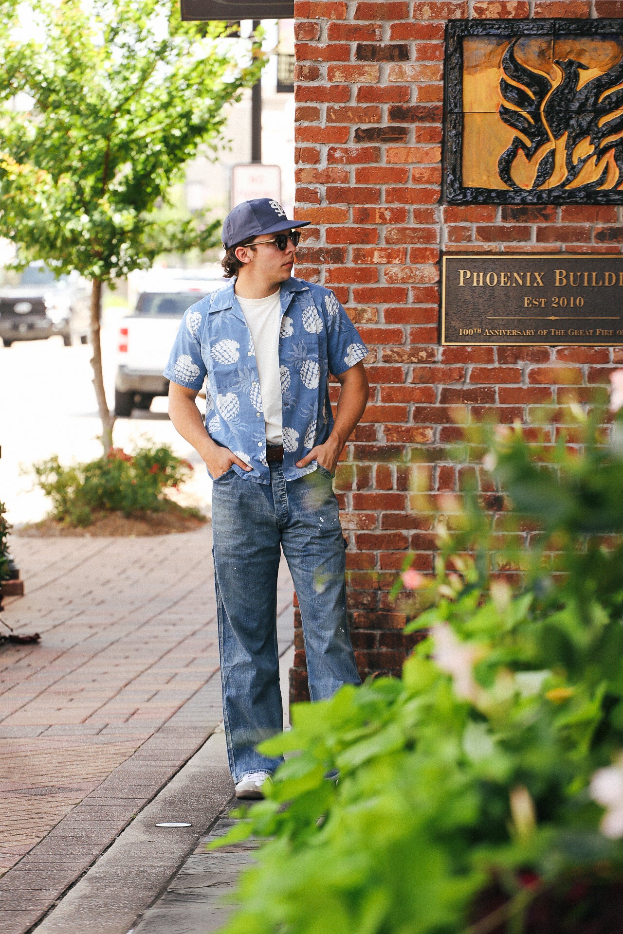 Man wearing indigo blue Hawaiian shirt with white pineapple print, jeans, and cap standing by brick wall plaque