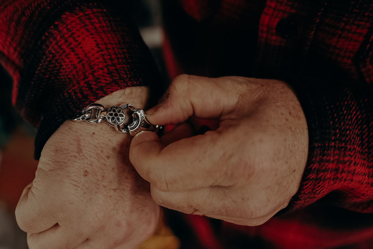 Close-up of hands fastening a silver chain bracelet with intricate flower design, wearing a red plaid shirt