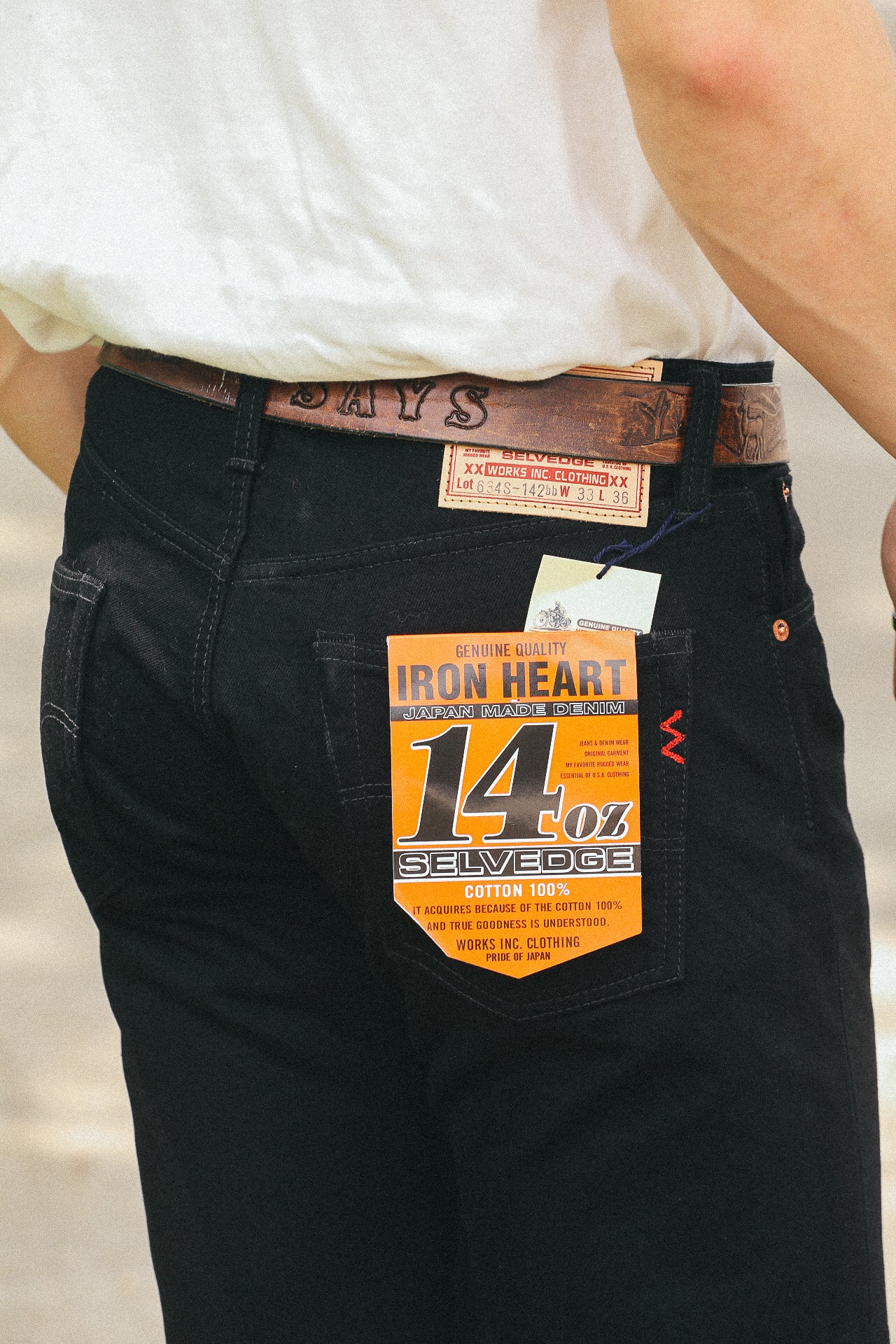 Close-up of person wearing black Iron Heart 14 oz selvedge denim jeans with leather belt and tags