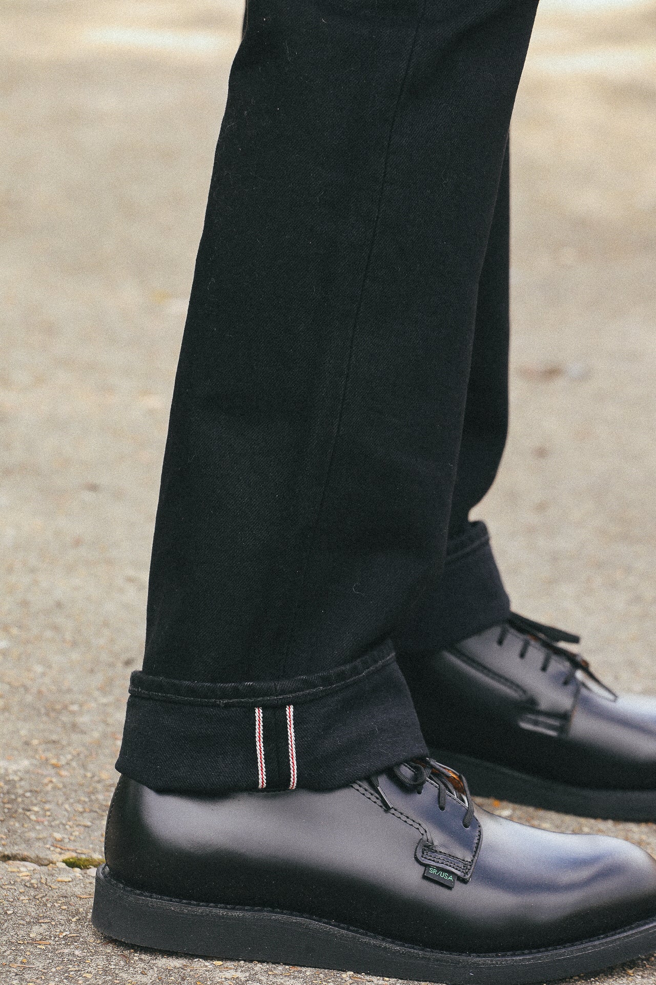 Close-up of black selvedge denim jeans cuff with red and white stitching paired with black polished leather shoes on concrete
