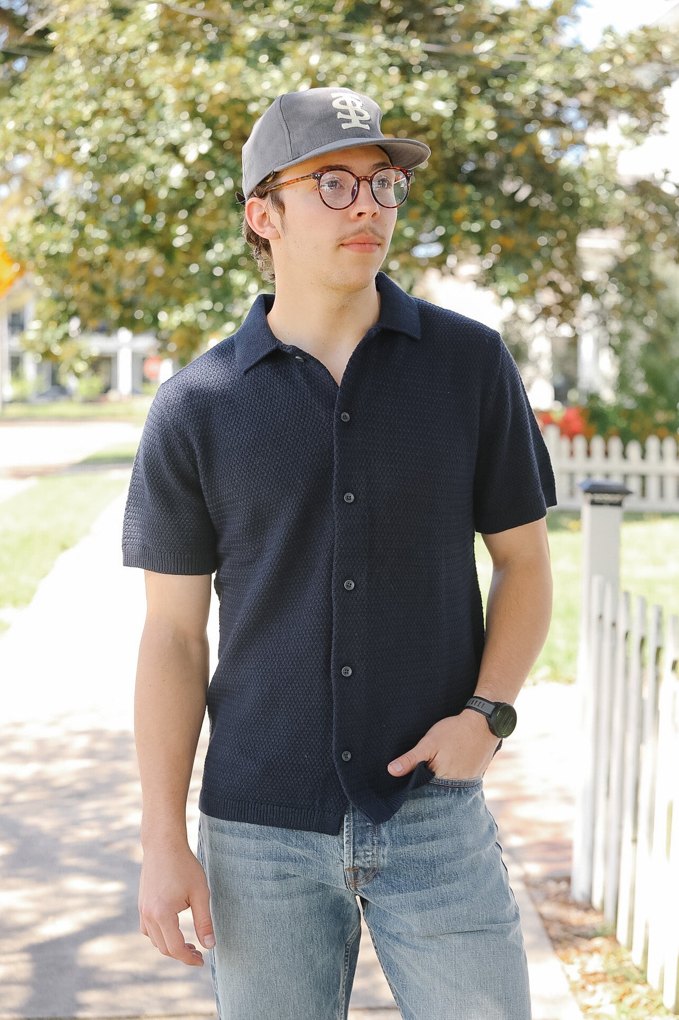Person wearing a dark shirt and cap outdoors with trees and a fence in the background