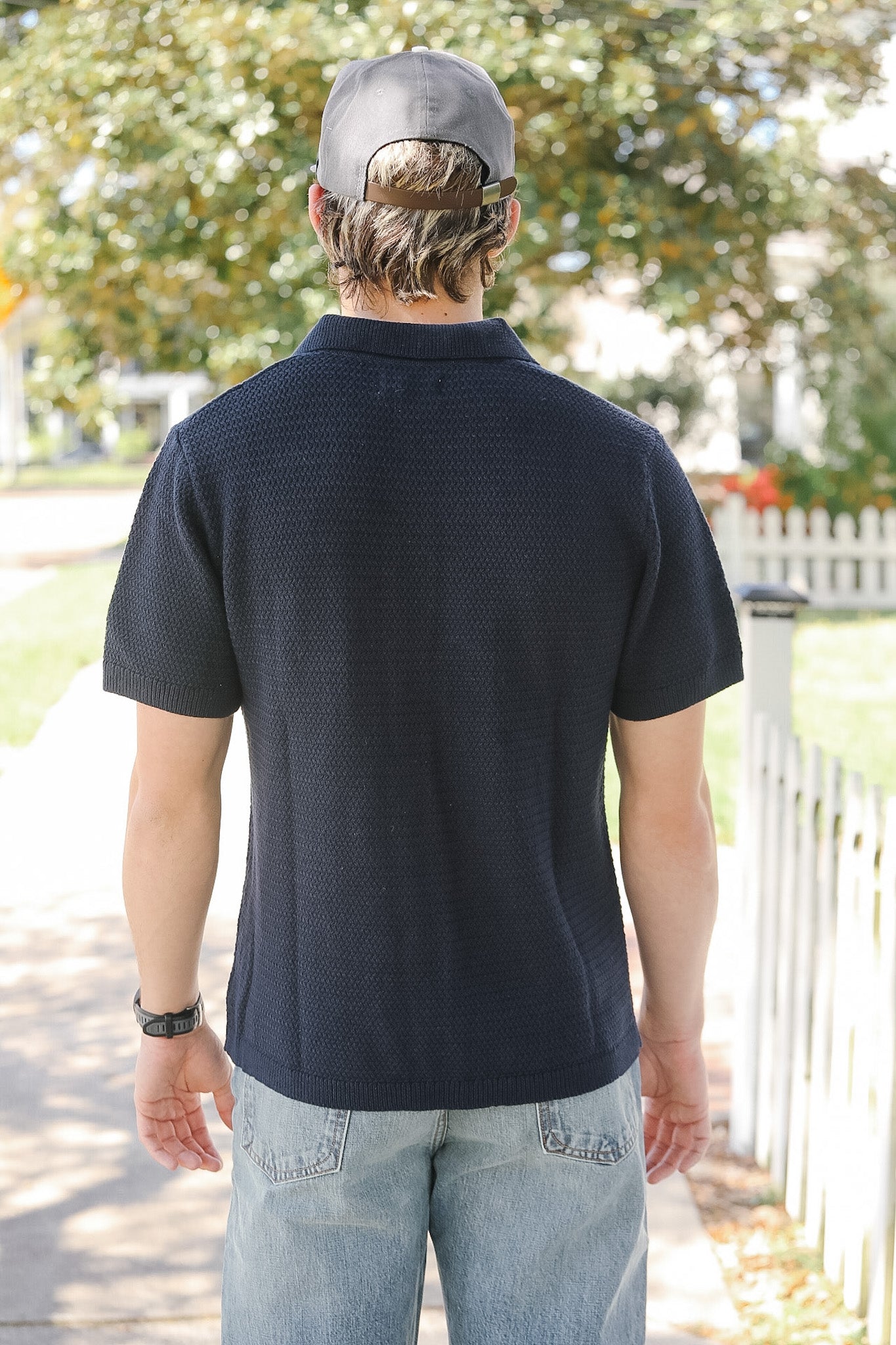 Man wearing a navy blue polo shirt and light blue jeans with a cap, standing outdoors.