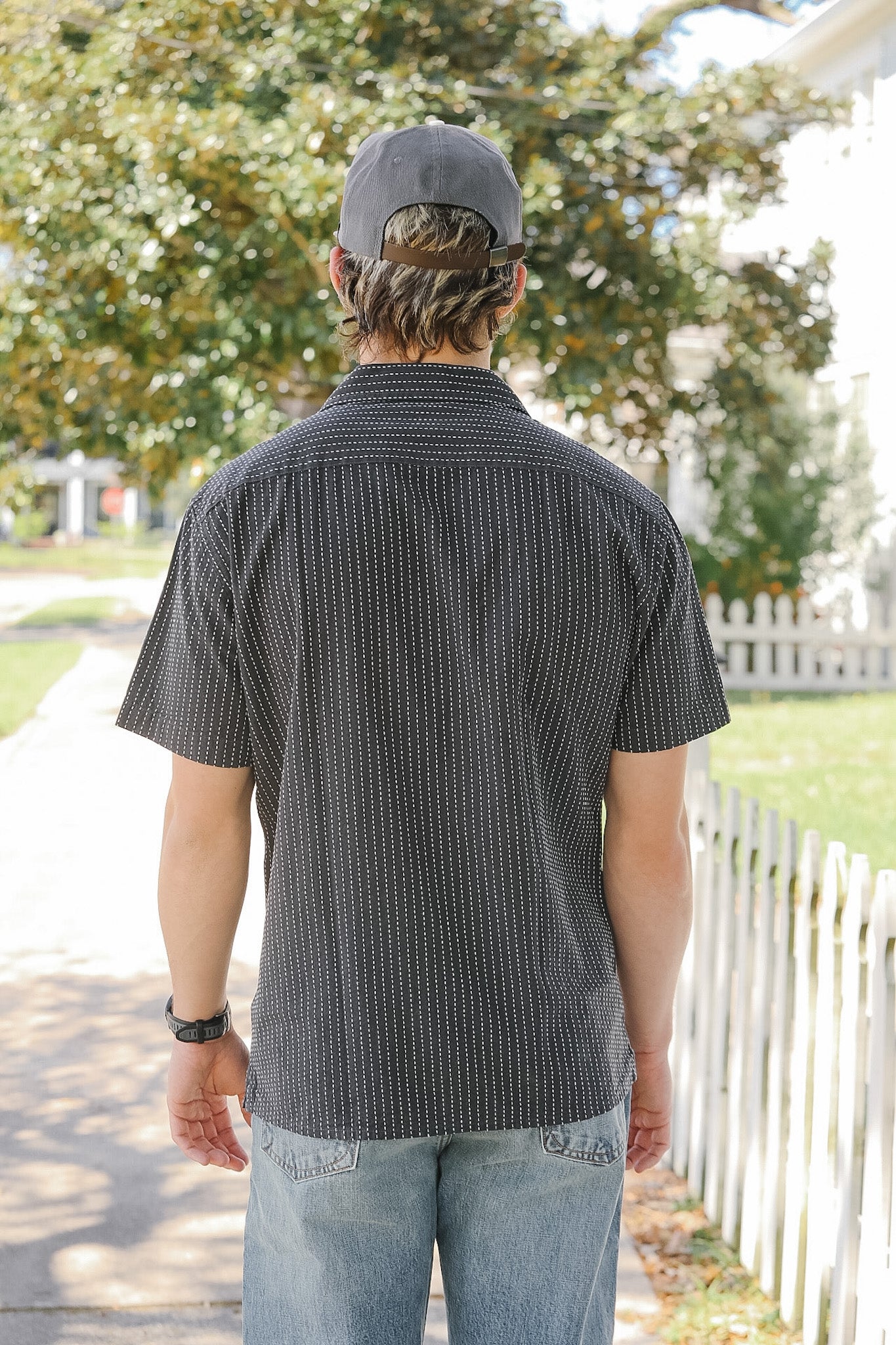 Person wearing a gray patterned shirt and cap, standing outdoors with trees and a white fence in the background.
