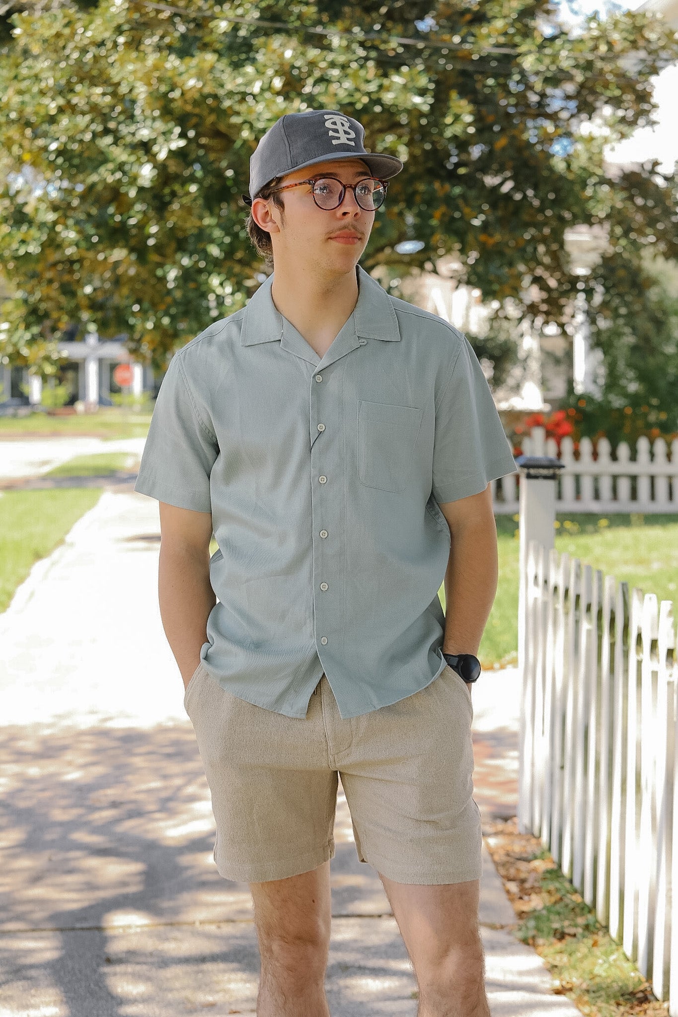 Man wearing a light gray button-up shirt and beige shorts standing on a sidewalk with trees and a white picket fence in the background.
