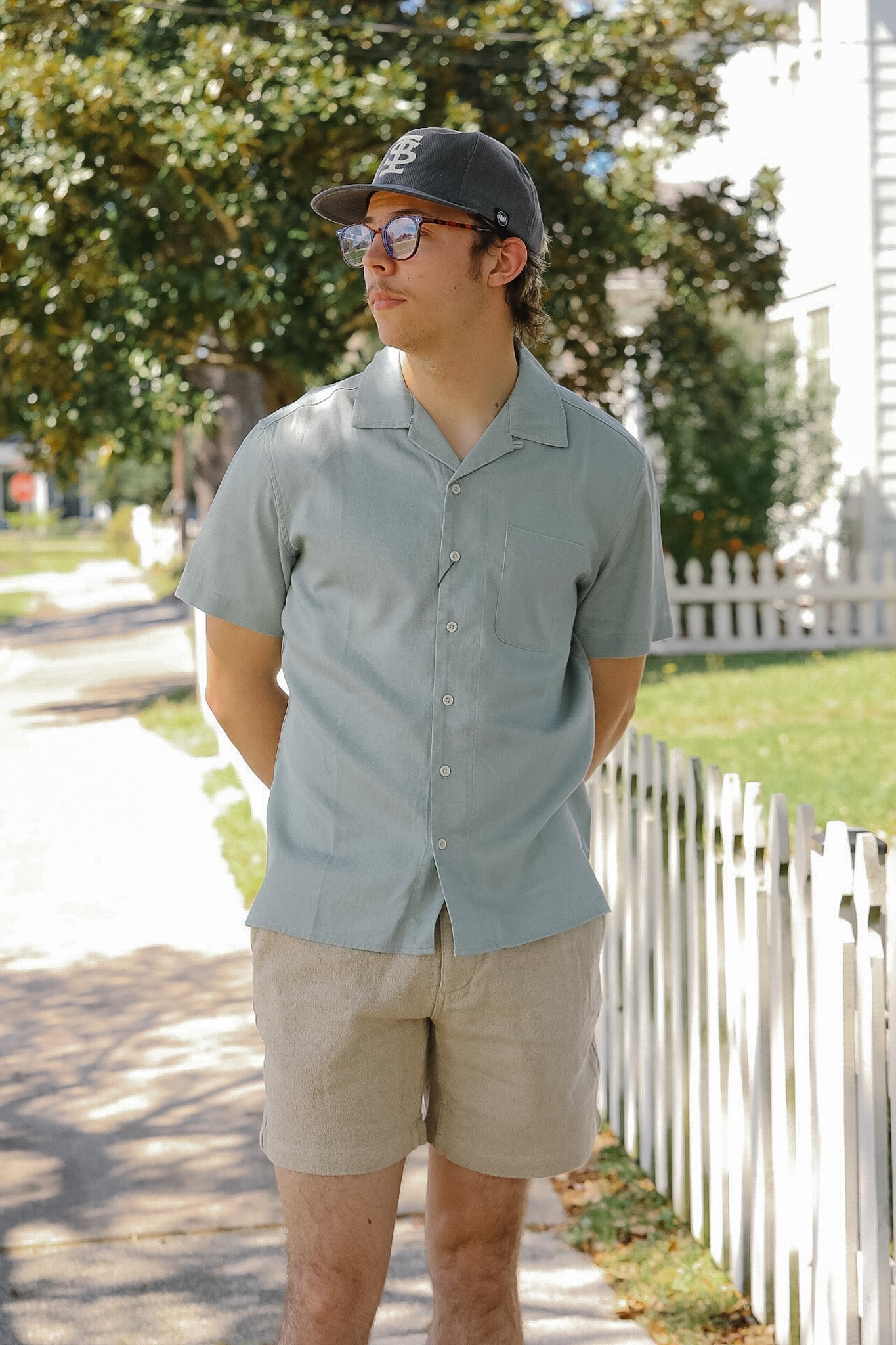 Man wearing a light gray shirt and beige shorts standing outdoors with trees and a white fence in the background.