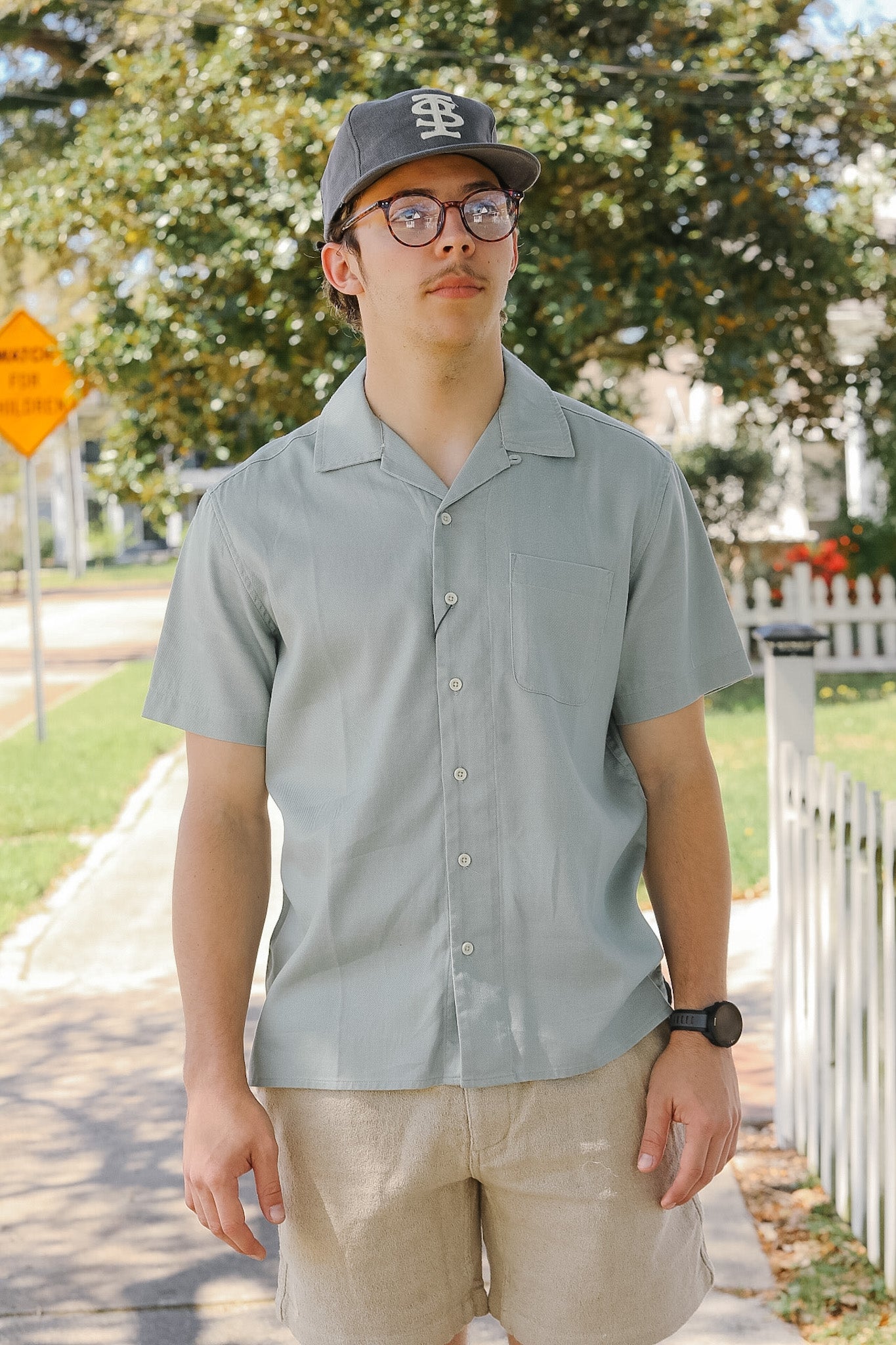 Person wearing a light gray shirt and cap outdoors with trees and a fence in the background