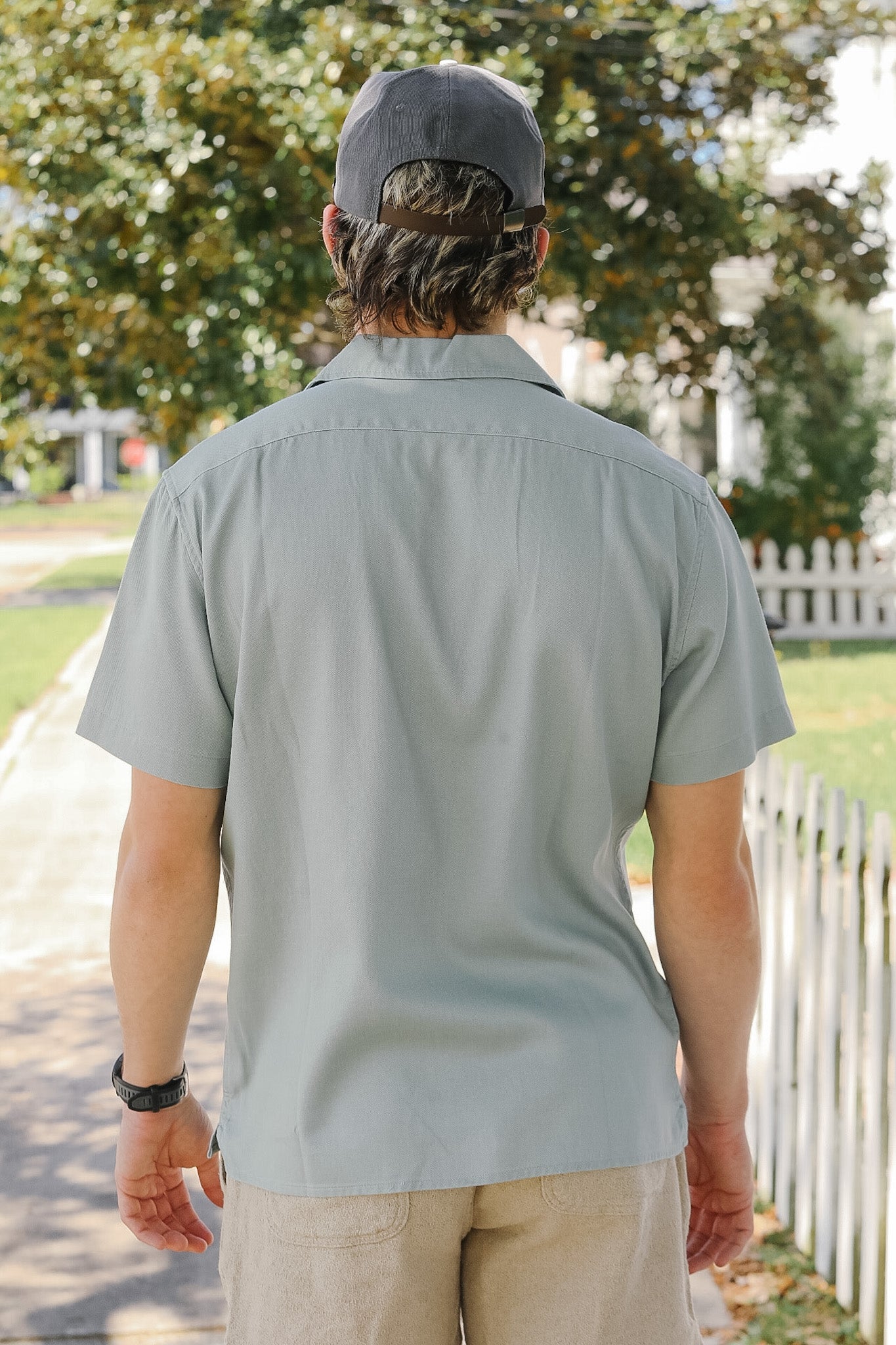 Person wearing a light gray shirt and cap walking outdoors with trees and a white picket fence in the background.