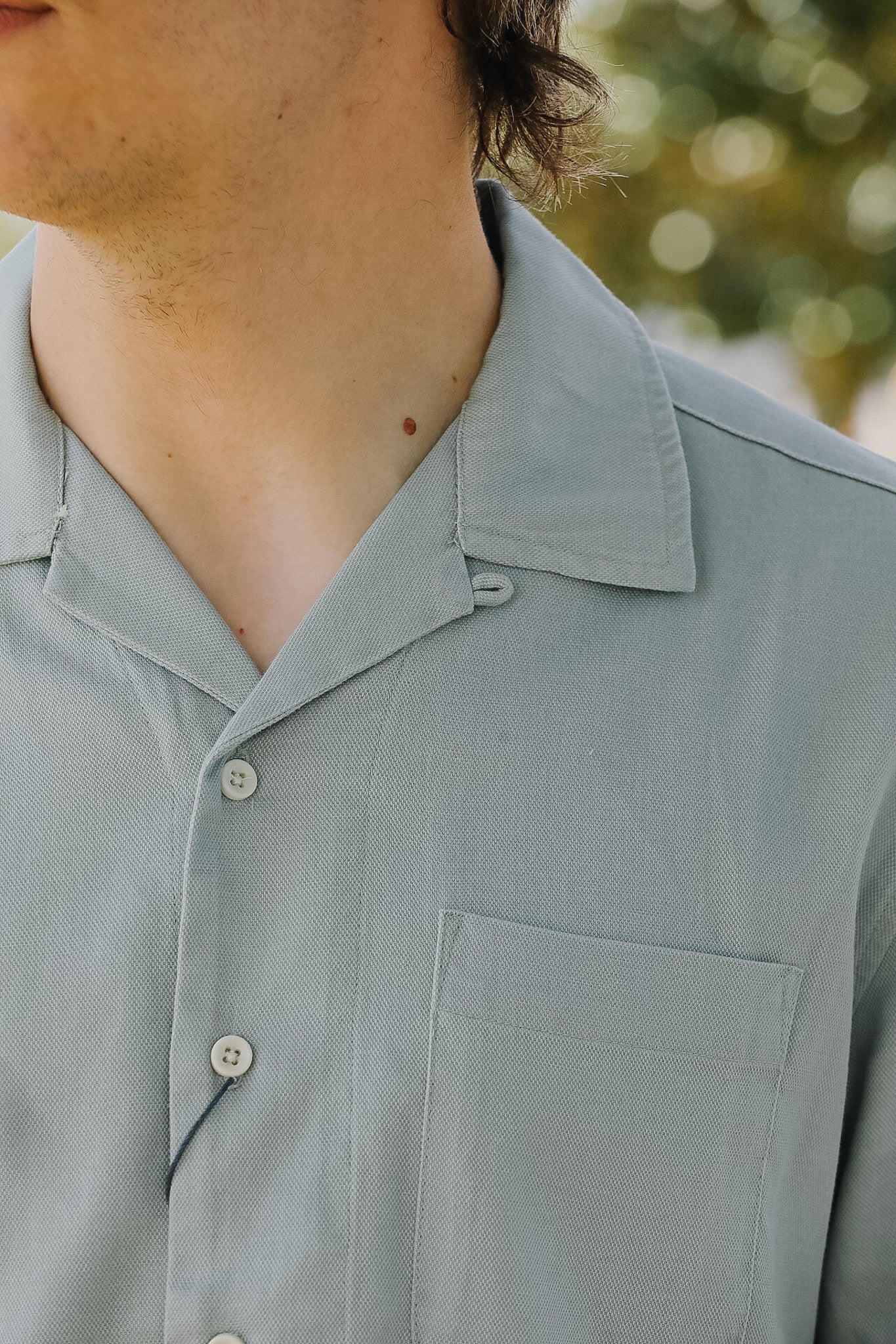 Close-up of a person wearing a light gray button-up shirt with a blurred natural background