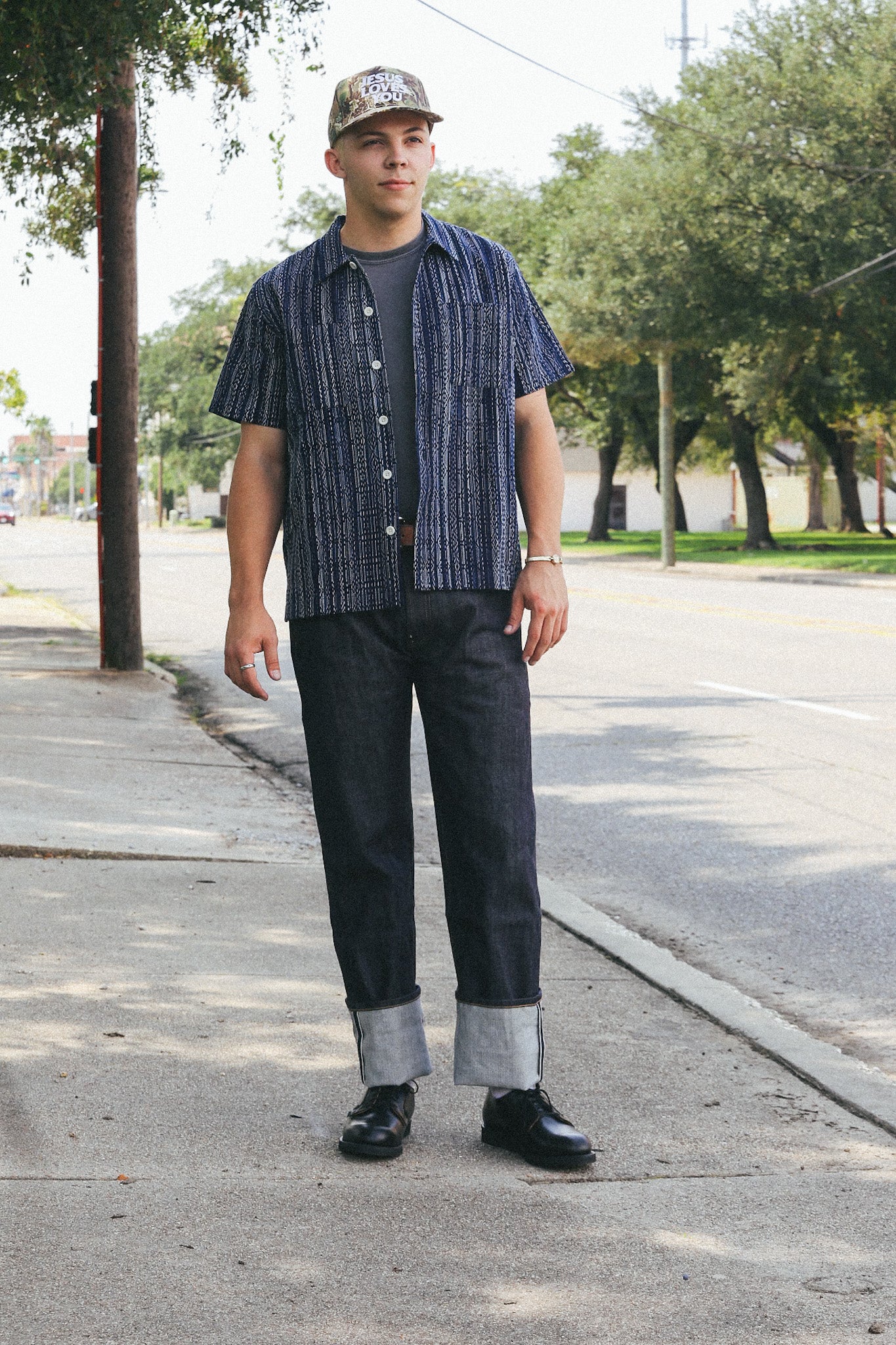Man wearing dark patterned short-sleeve shirt, cuffed dark jeans, black shoes, and camo cap standing on sidewalk