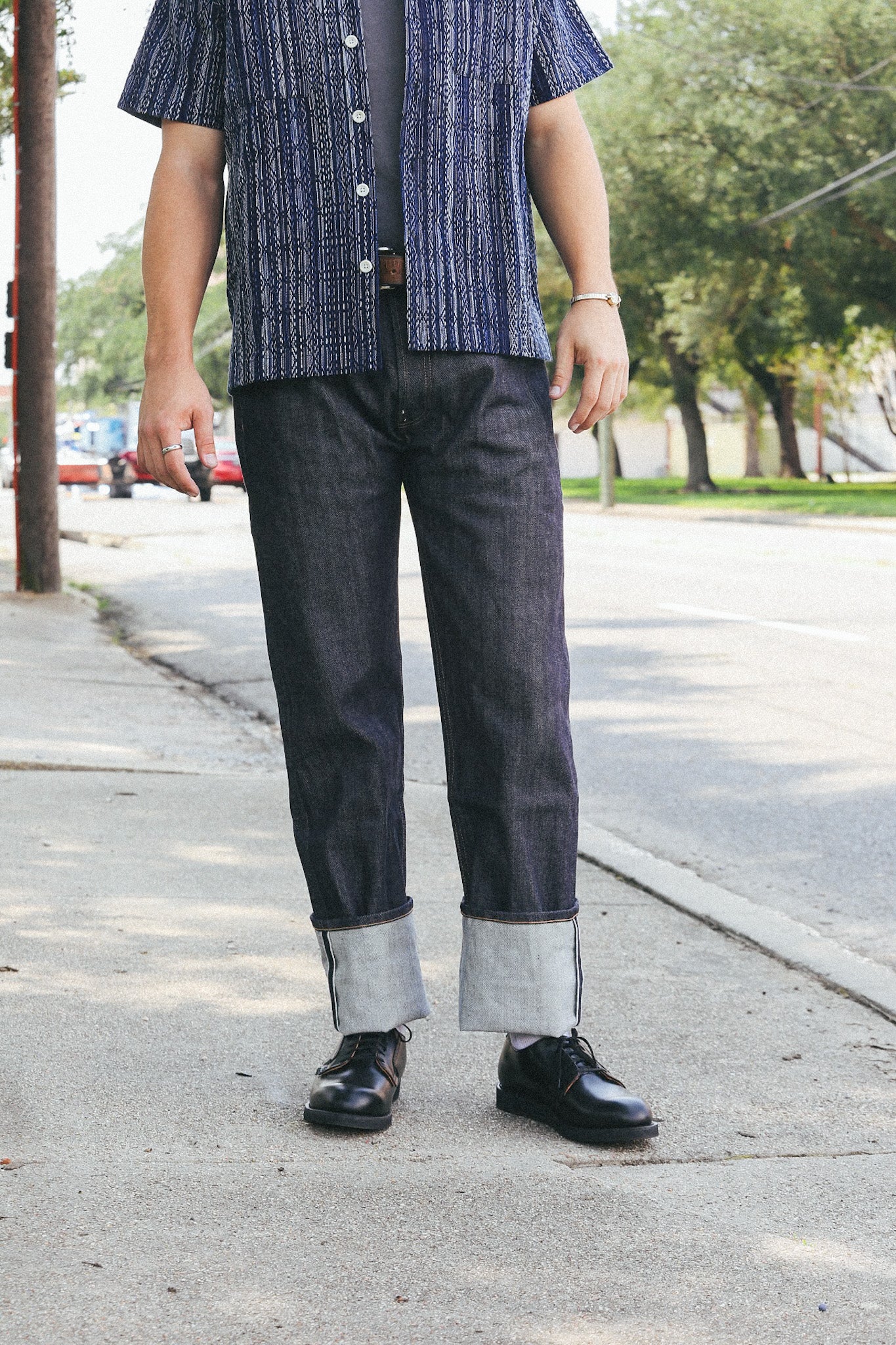Casual male outfit with cuffed dark denim jeans, black leather shoes, and a patterned short sleeve shirt on city sidewalk