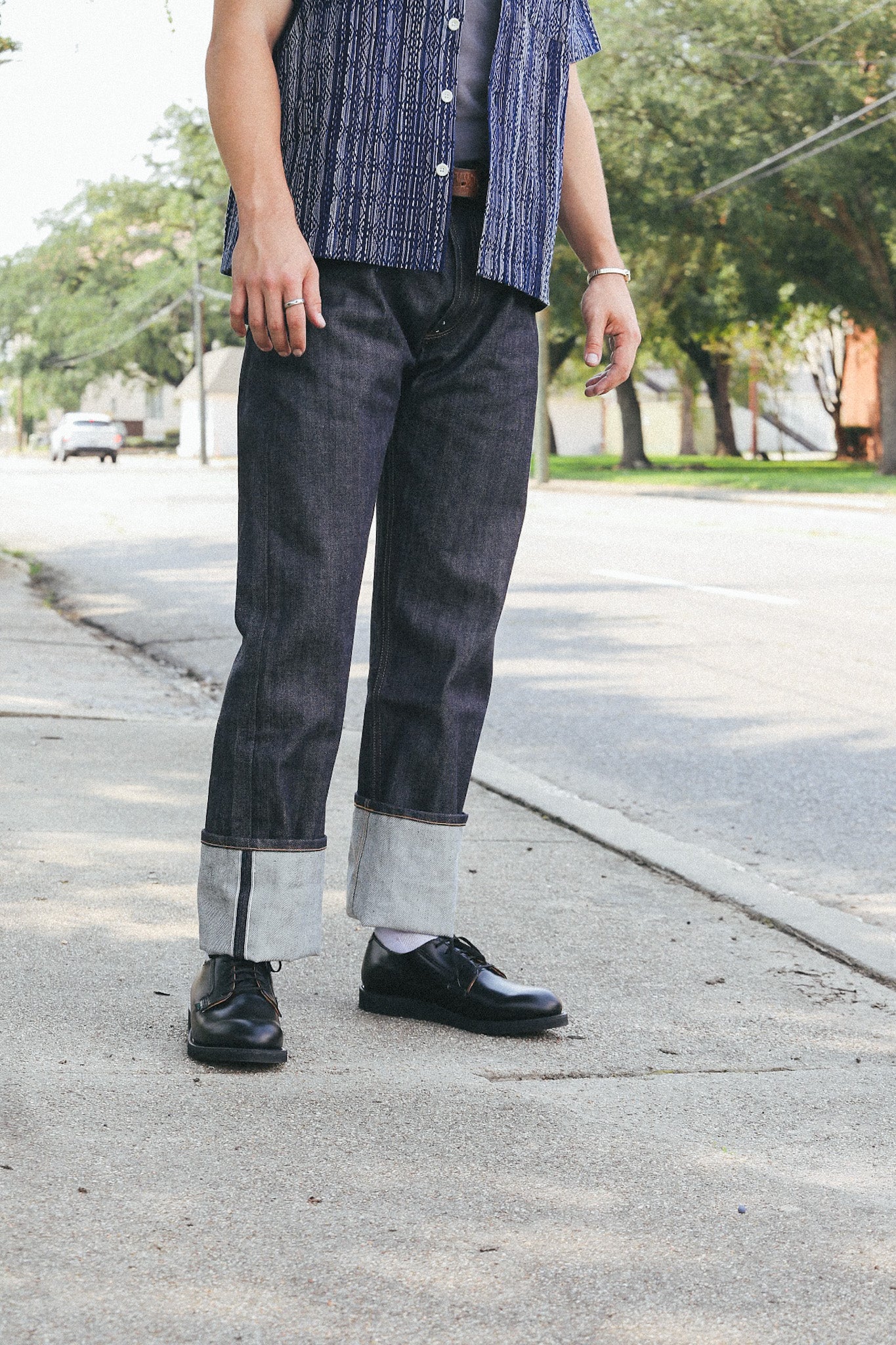 Person standing on sidewalk wearing cuffed dark denim jeans, black leather shoes, and a patterned blue shirt