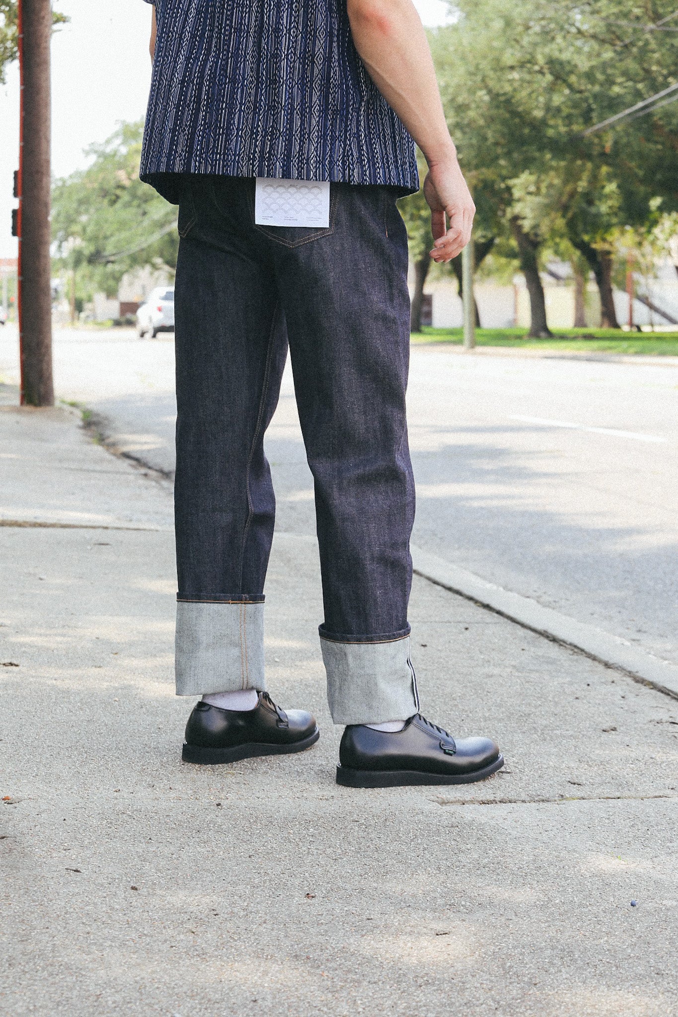 Man wearing dark raw denim jeans with large cuffs and black leather shoes standing on sidewalk