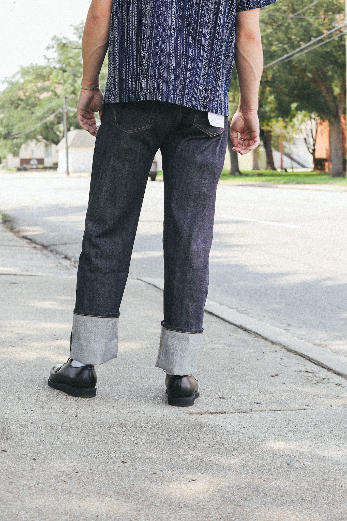 Rear view of person wearing rolled cuff dark denim jeans and black leather shoes on sidewalk