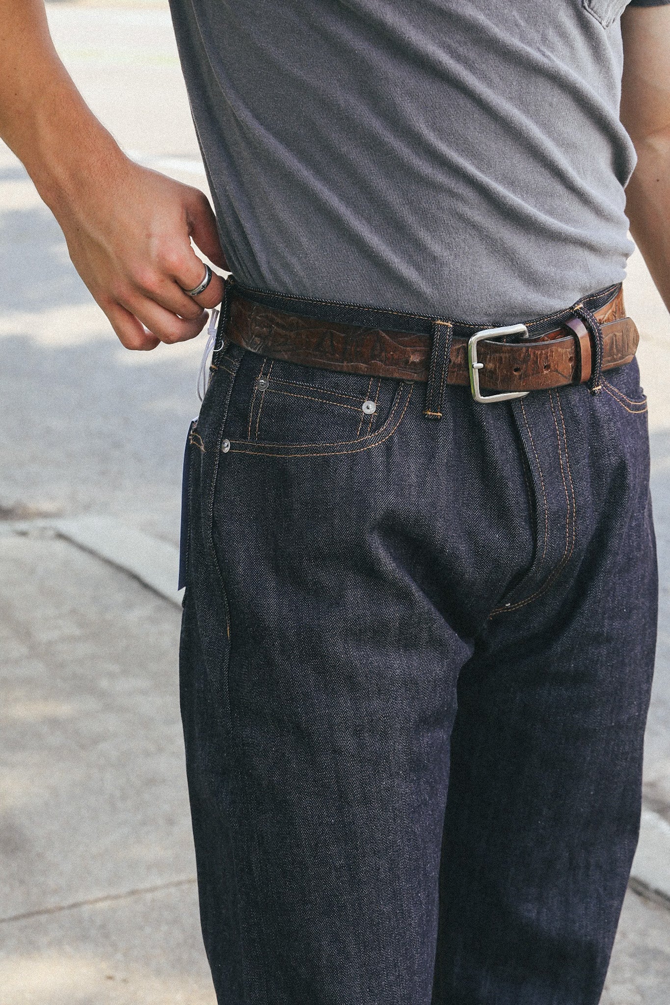 Close-up of person wearing dark denim jeans and a textured brown leather belt with a silver buckle