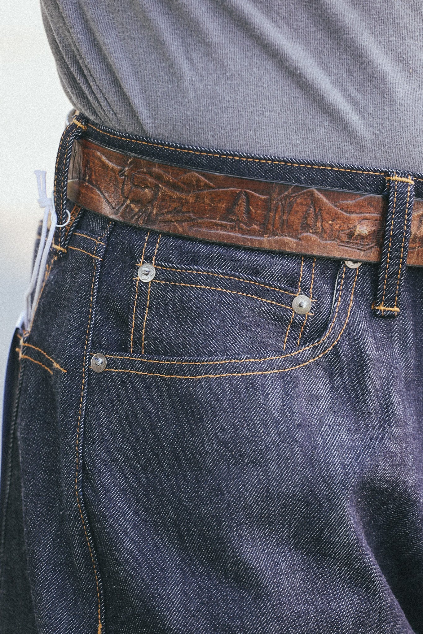 Close-up of dark blue denim jeans with orange stitching and a brown leather belt featuring carved nature scenes