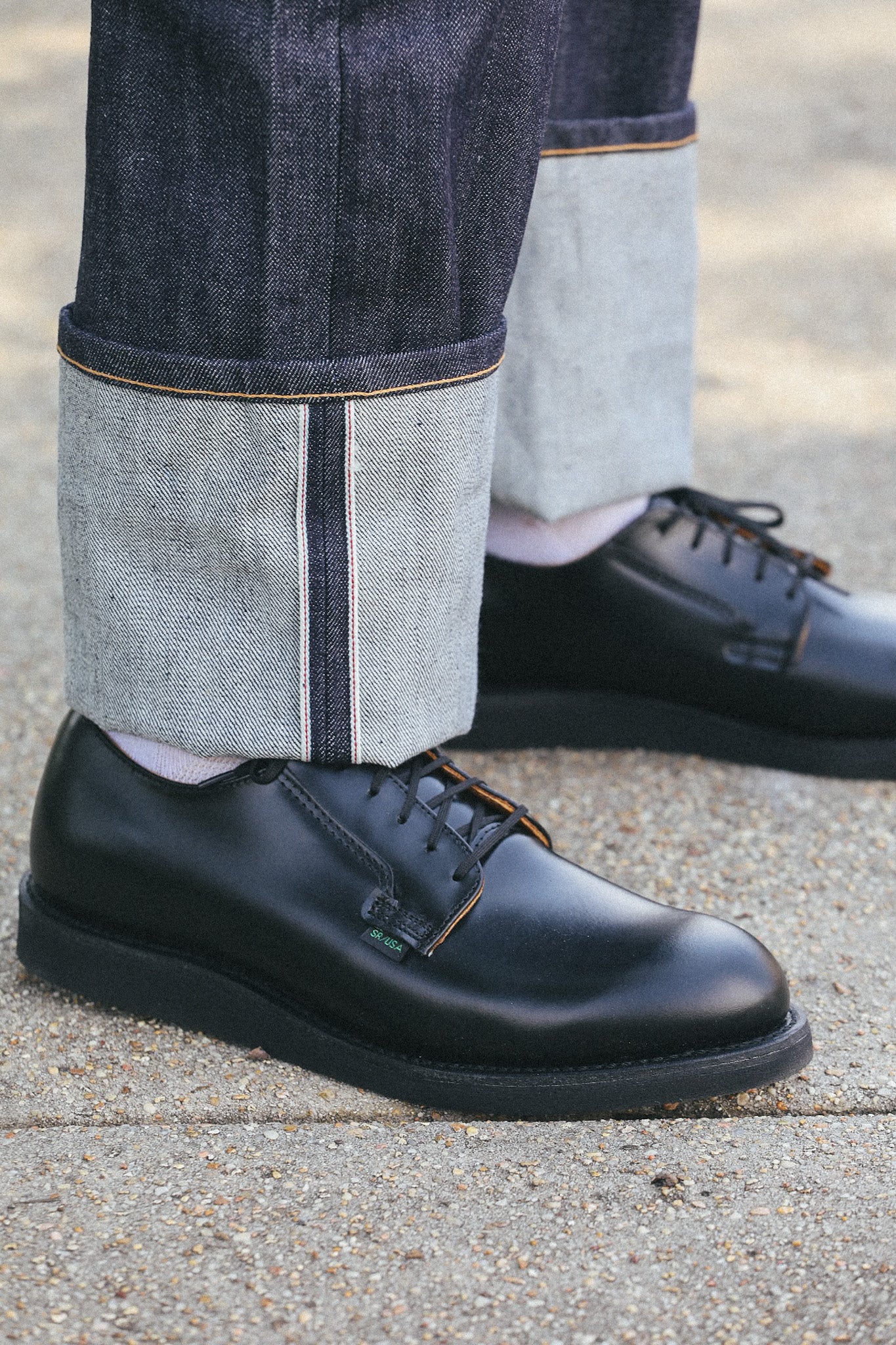 Close-up of black leather SR USA shoes paired with rolled-up selvedge denim jeans on concrete