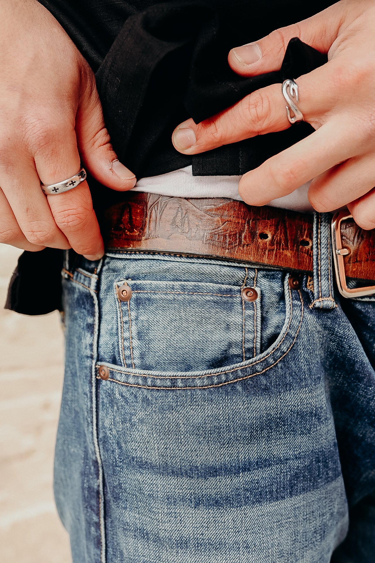 Close-up of hands wearing silver rings adjusting embossed brown leather belt with blue denim jeans