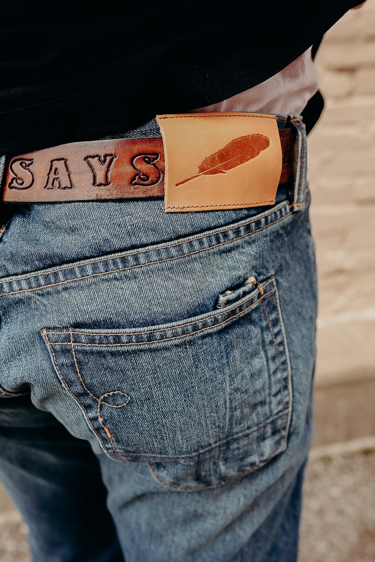 Close-up of person wearing blue denim jeans with a brown leather belt featuring embossed letters and a feather emblem