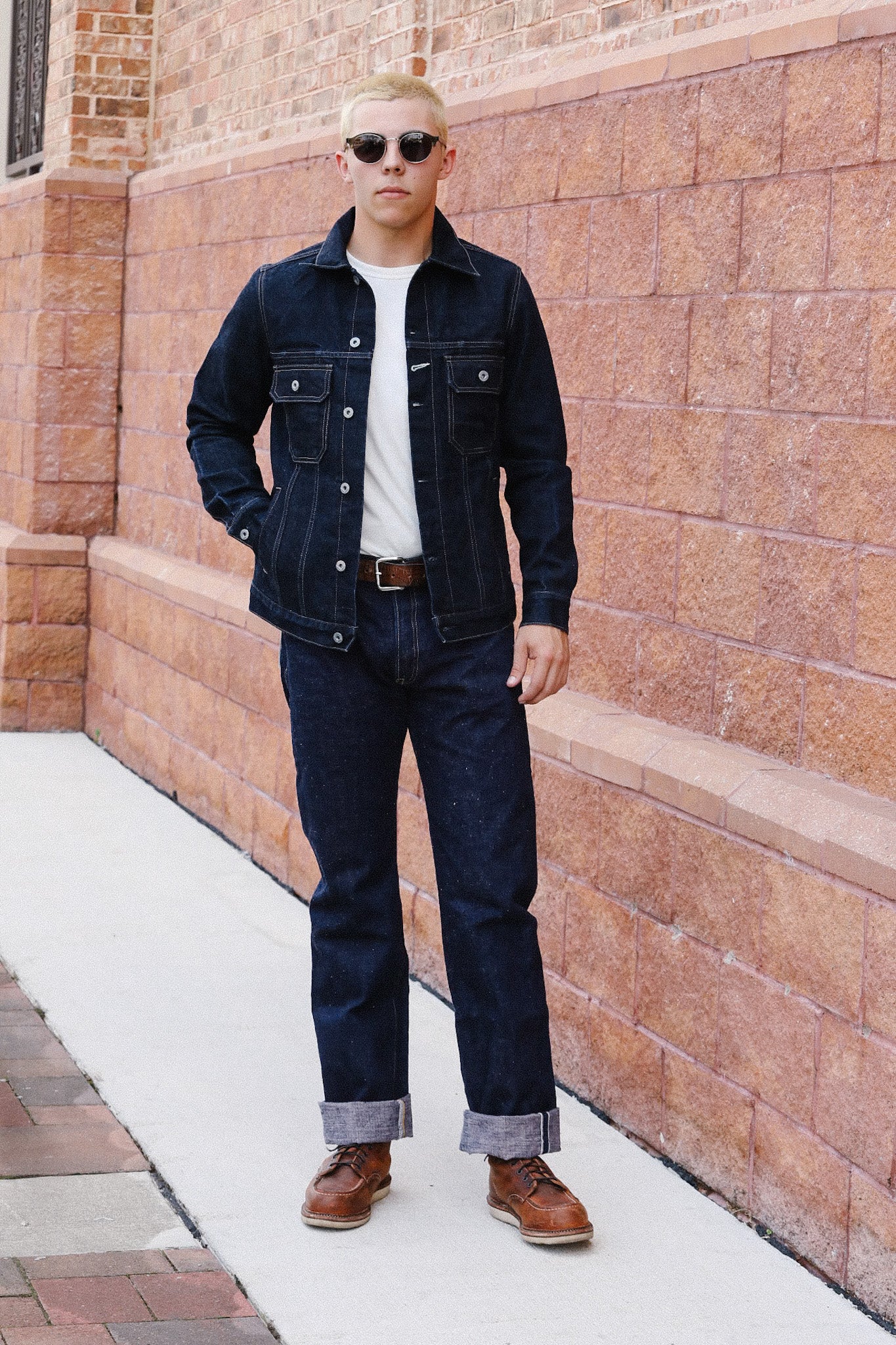man wearing dark denim jacket and jeans with brown leather boots standing on sidewalk by brick wall