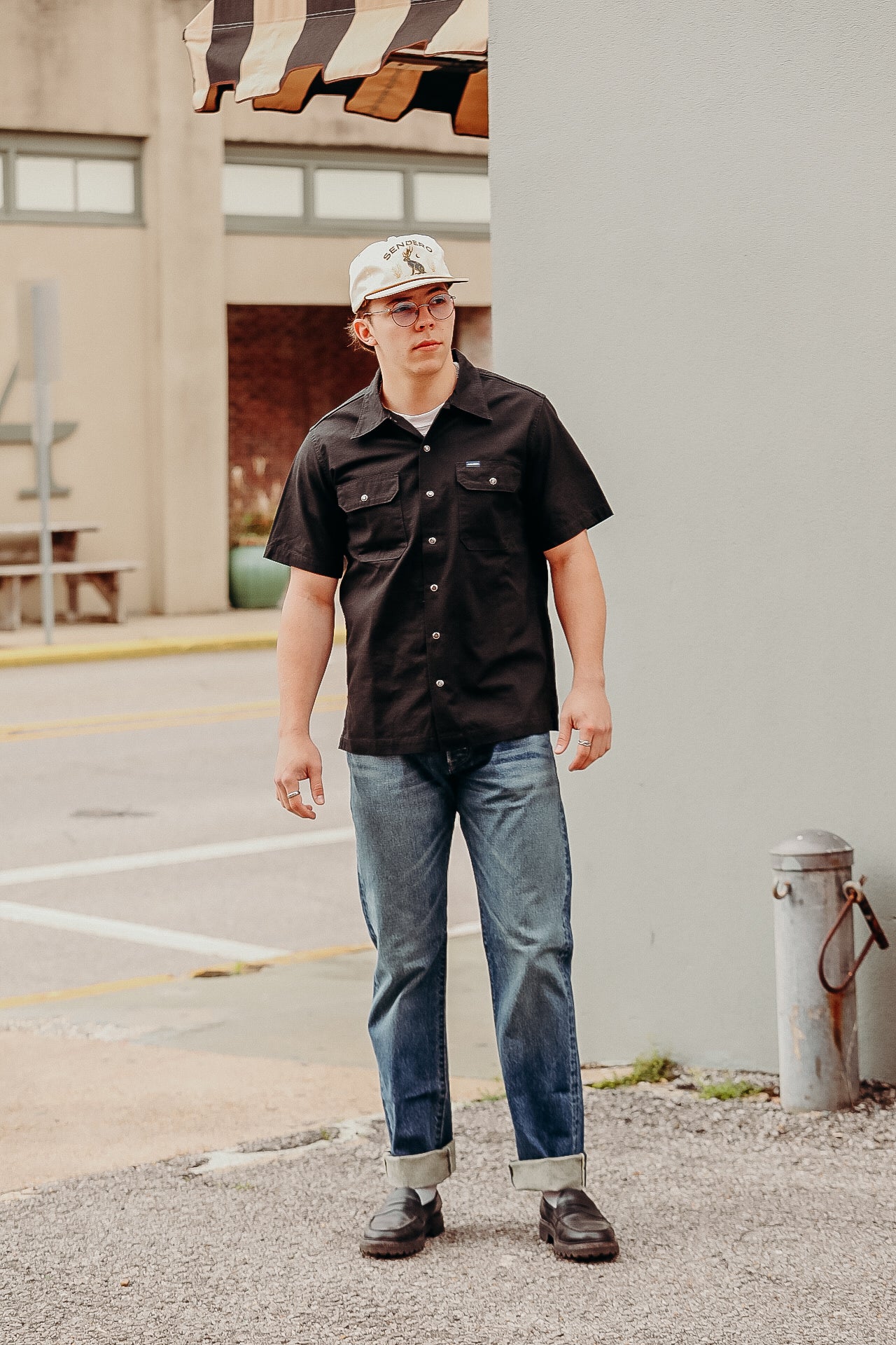 Young man wearing black work shirt, jeans, and white cap standing outside near building under brown striped awning