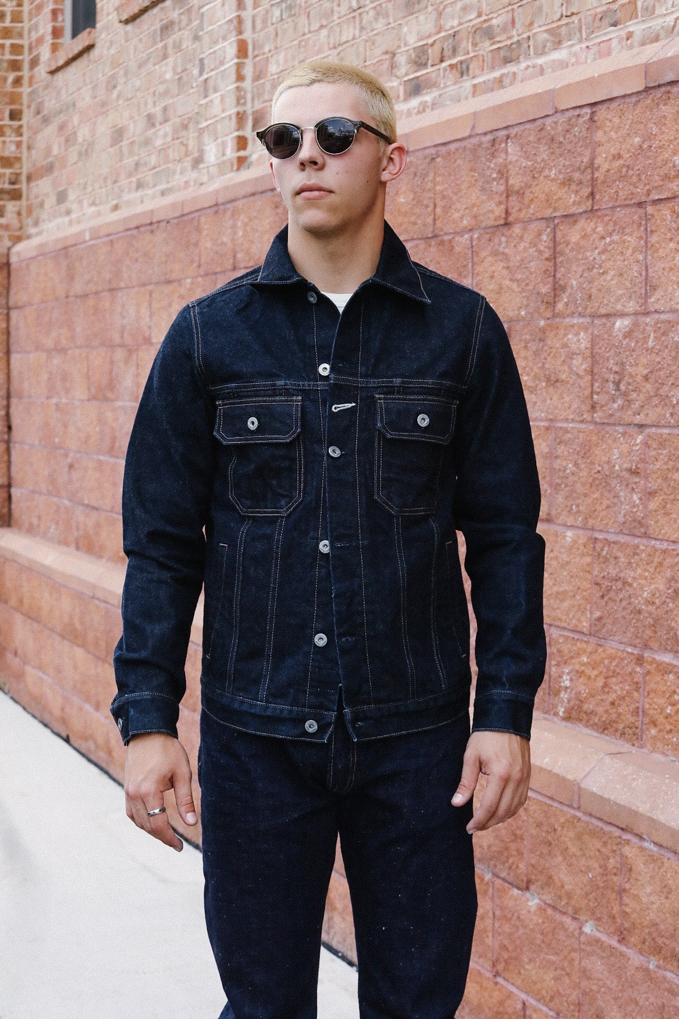 Man wearing black denim jacket and pants standing by brick wall wearing sunglasses