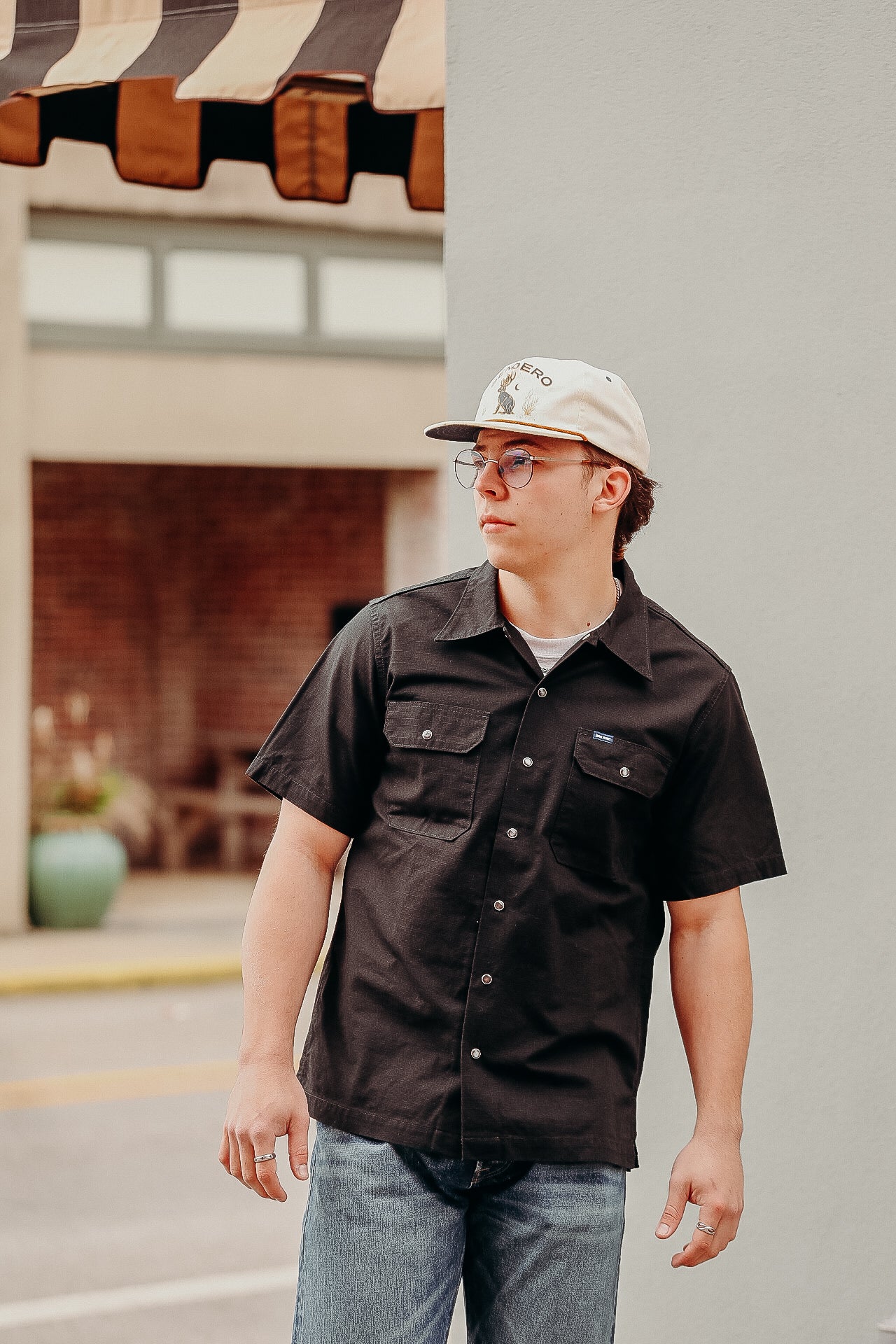 Young man wearing black short-sleeve button shirt and beige cap standing outdoors near building