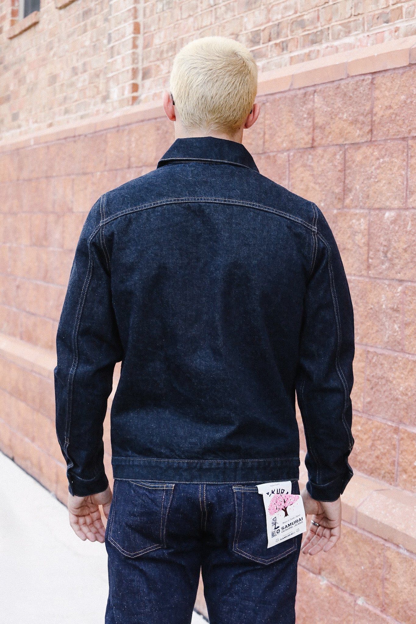 Man with blonde hair wearing dark denim jacket and jeans with Samurai brand tag, standing by brick wall
