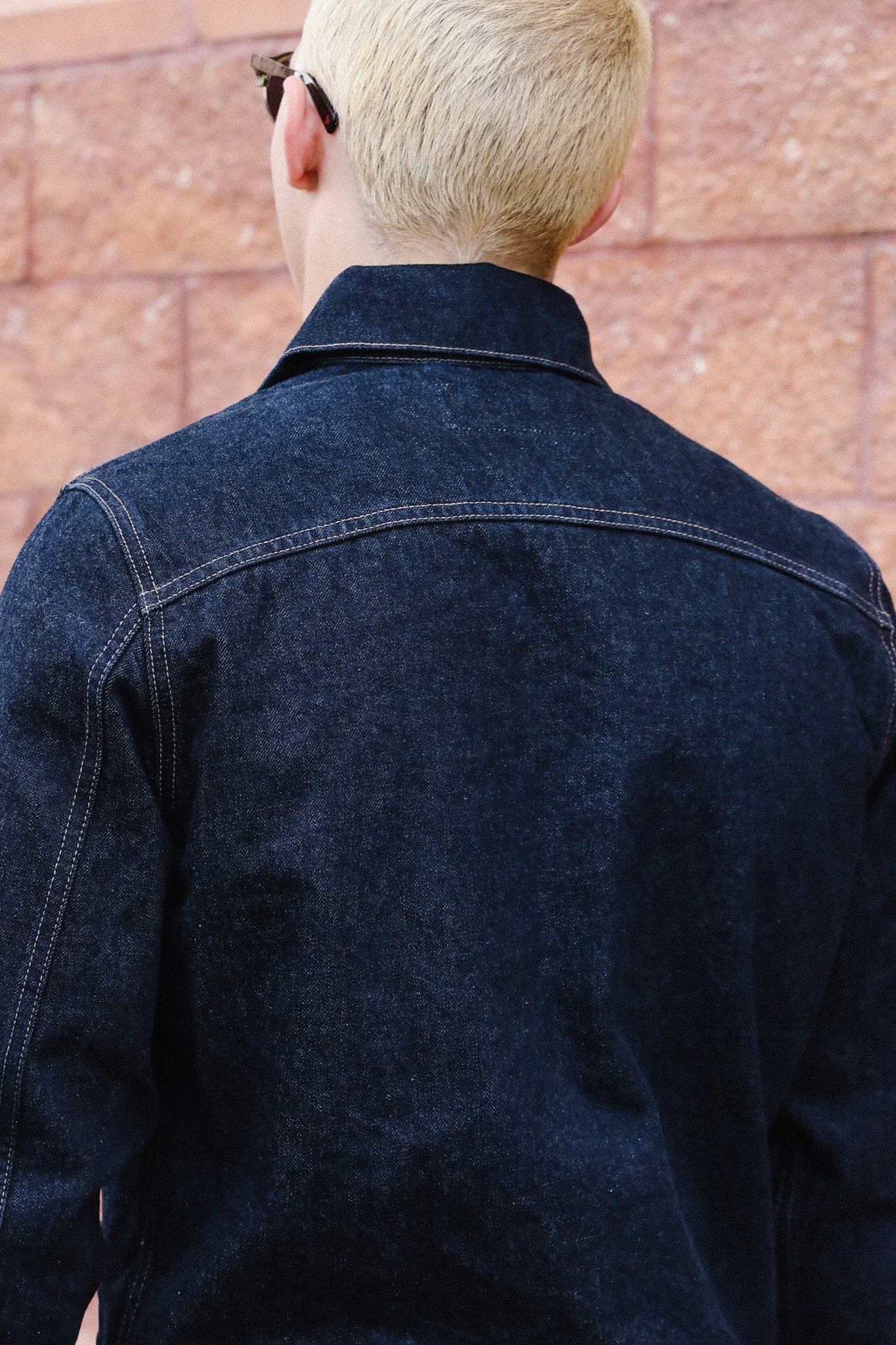 Back view of person with blonde hair wearing dark denim jacket and black sunglasses against brick wall