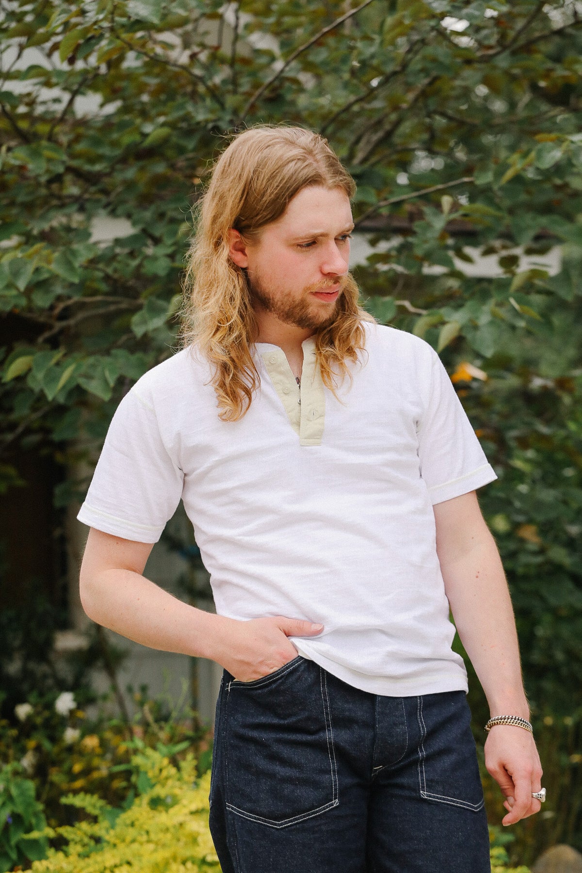 Man with long hair wearing a white shirt and dark pants standing outdoors with greenery in the background