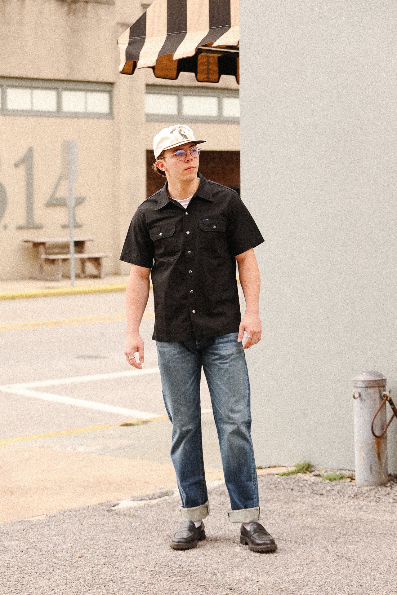 Man wearing black work shirt, rolled jeans, loafers, and white cap standing on urban street