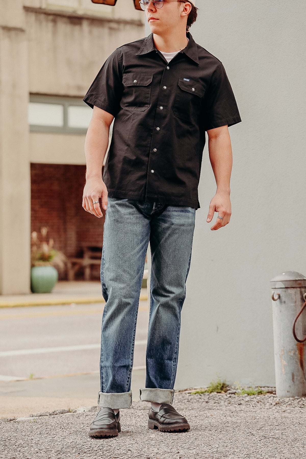 Man wearing black short-sleeve button-up work shirt and rolled-up blue jeans on urban street