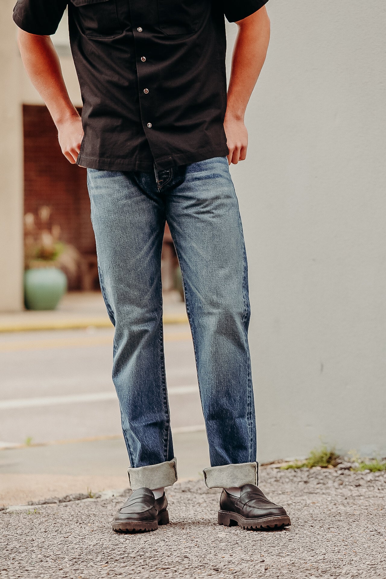 Person wearing cuffed blue jeans, black button-up shirt, and black leather loafers standing outdoors