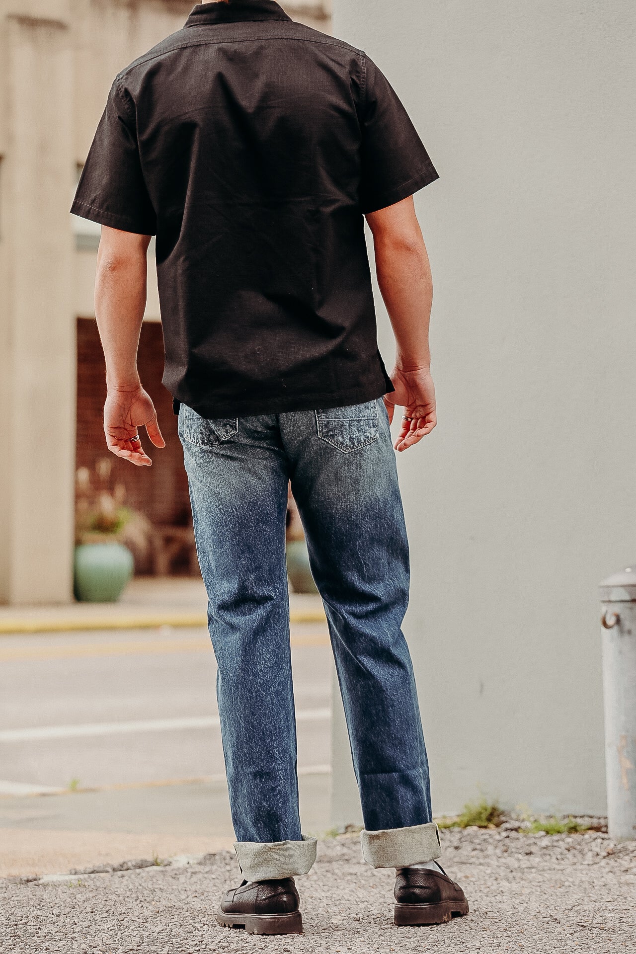Man wearing rolled-up blue jeans, black short-sleeve shirt, and black leather shoes outdoors