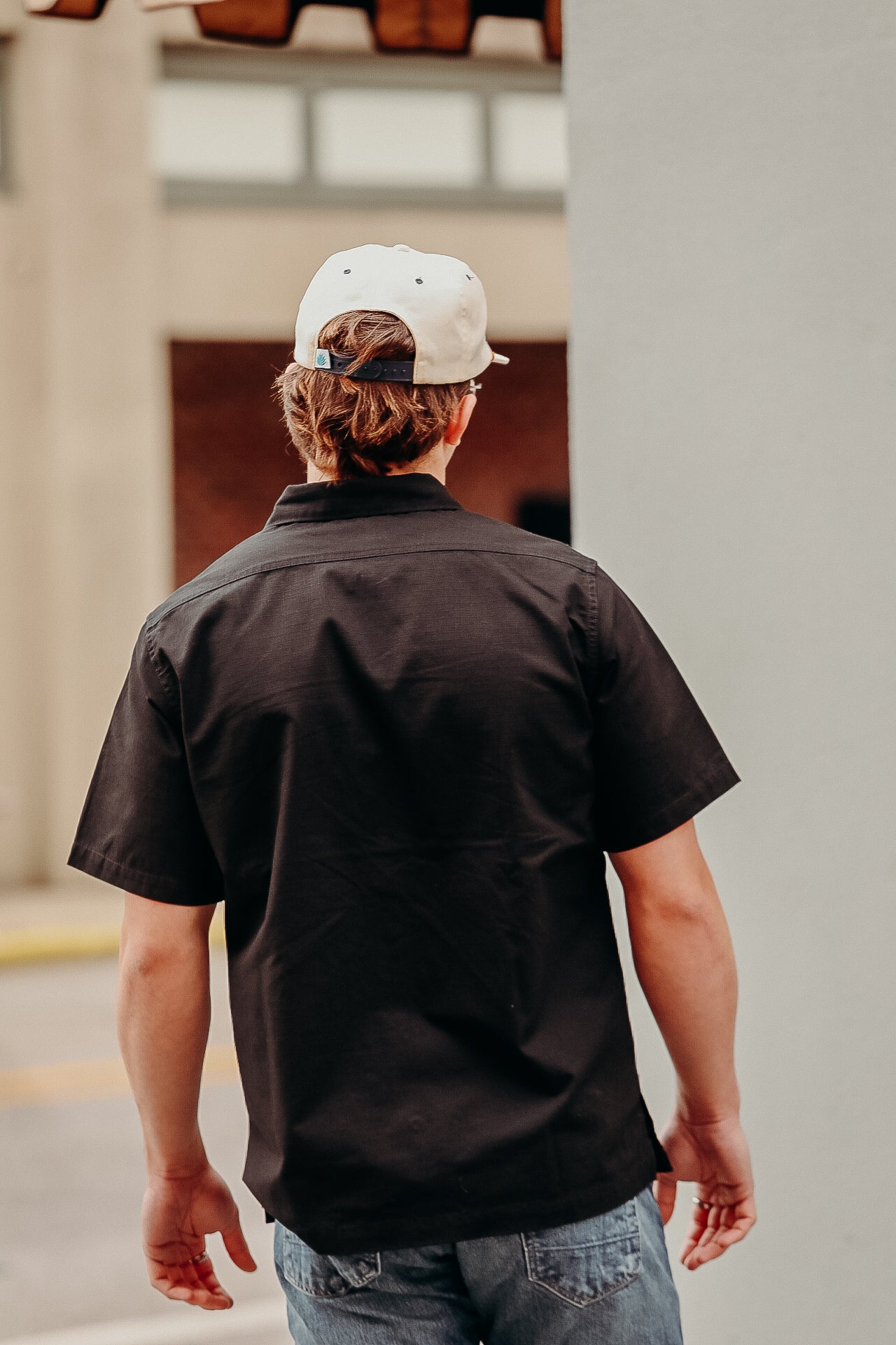 Man wearing black short-sleeve work shirt and white baseball cap standing outdoors, back view