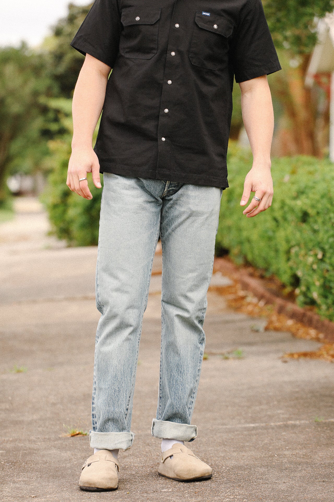 Man wearing black short-sleeve shirt with pockets, light blue rolled jeans, and beige footwear outdoors