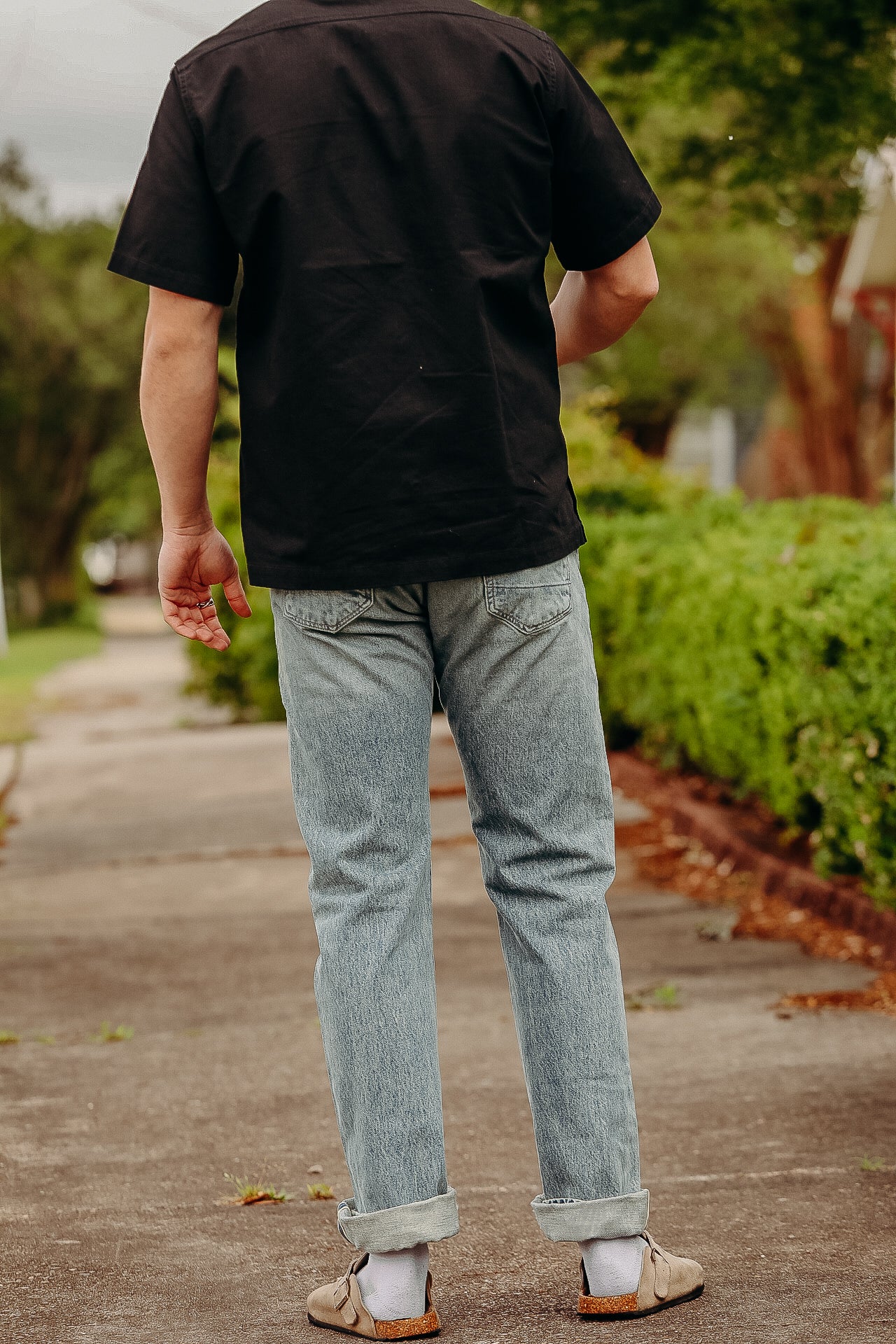 Rear view of a person wearing light blue denim jeans, black short-sleeve shirt, and beige sandals with socks on a sidewalk