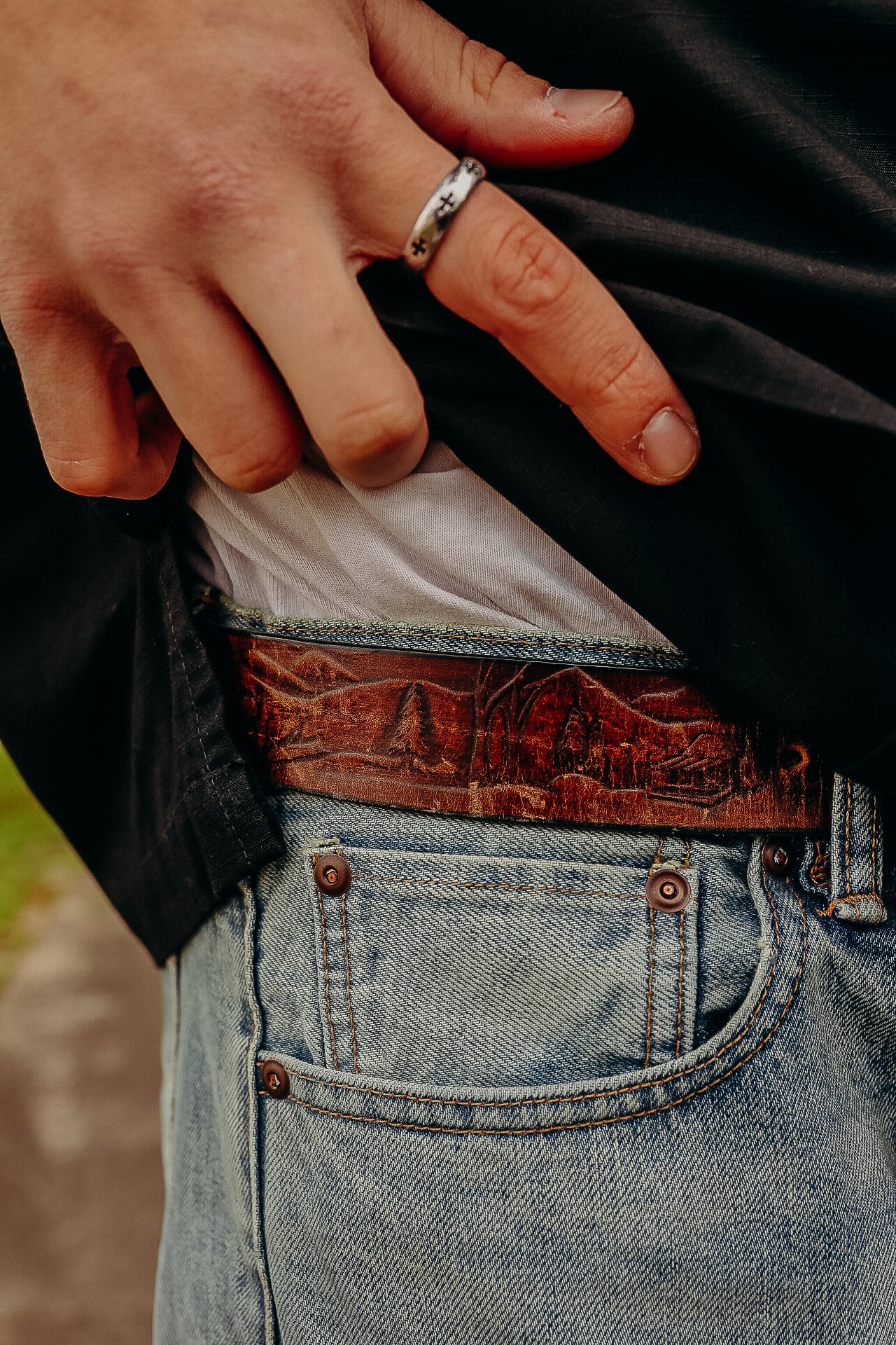 Hand wearing silver ring lifting black shirt to reveal embossed brown leather belt and blue denim jeans pocket