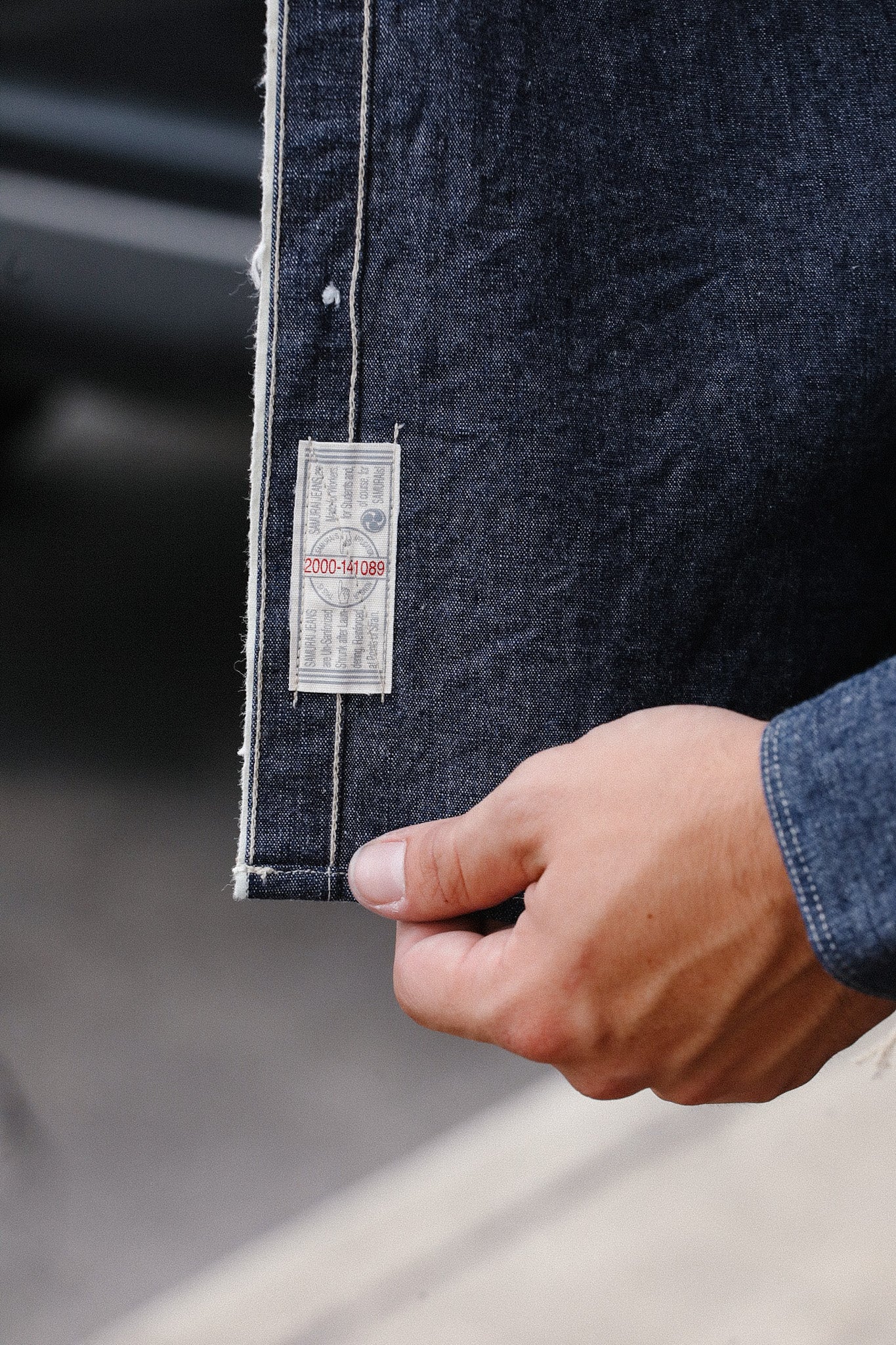 Hand holding dark denim fabric with visible white seam and product tag showing serial number at Iron Shop Provisions