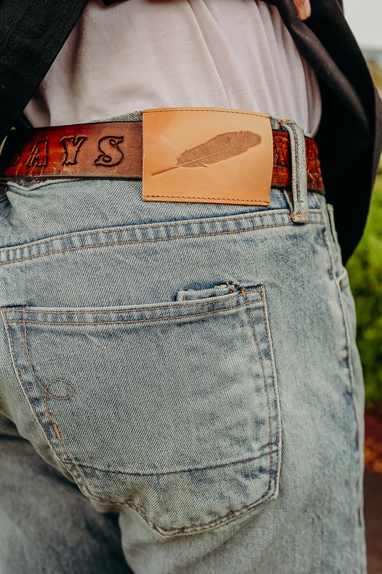 Close-up of light blue denim jeans with a brown leather belt and a tan leather patch featuring a feather design