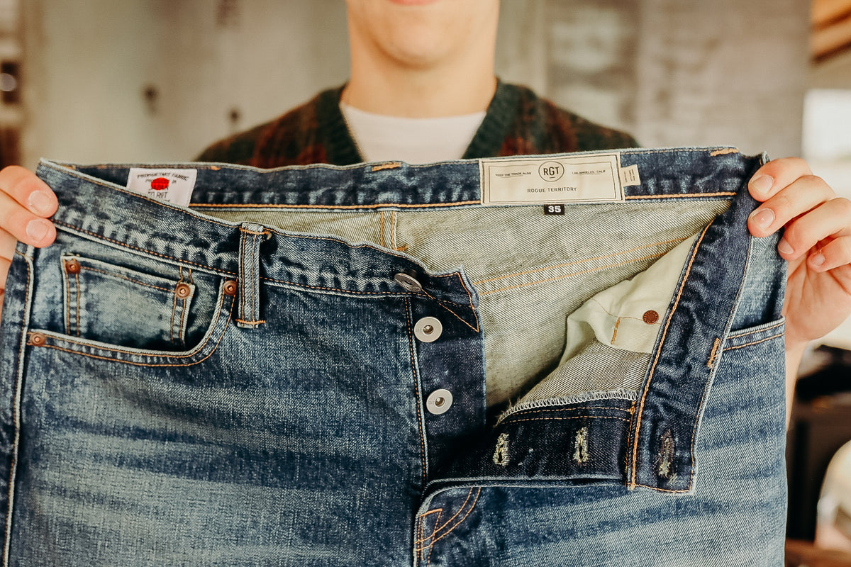 Person holding a pair of dark blue Rogue Territory denim jeans with button fly and visible inside tags