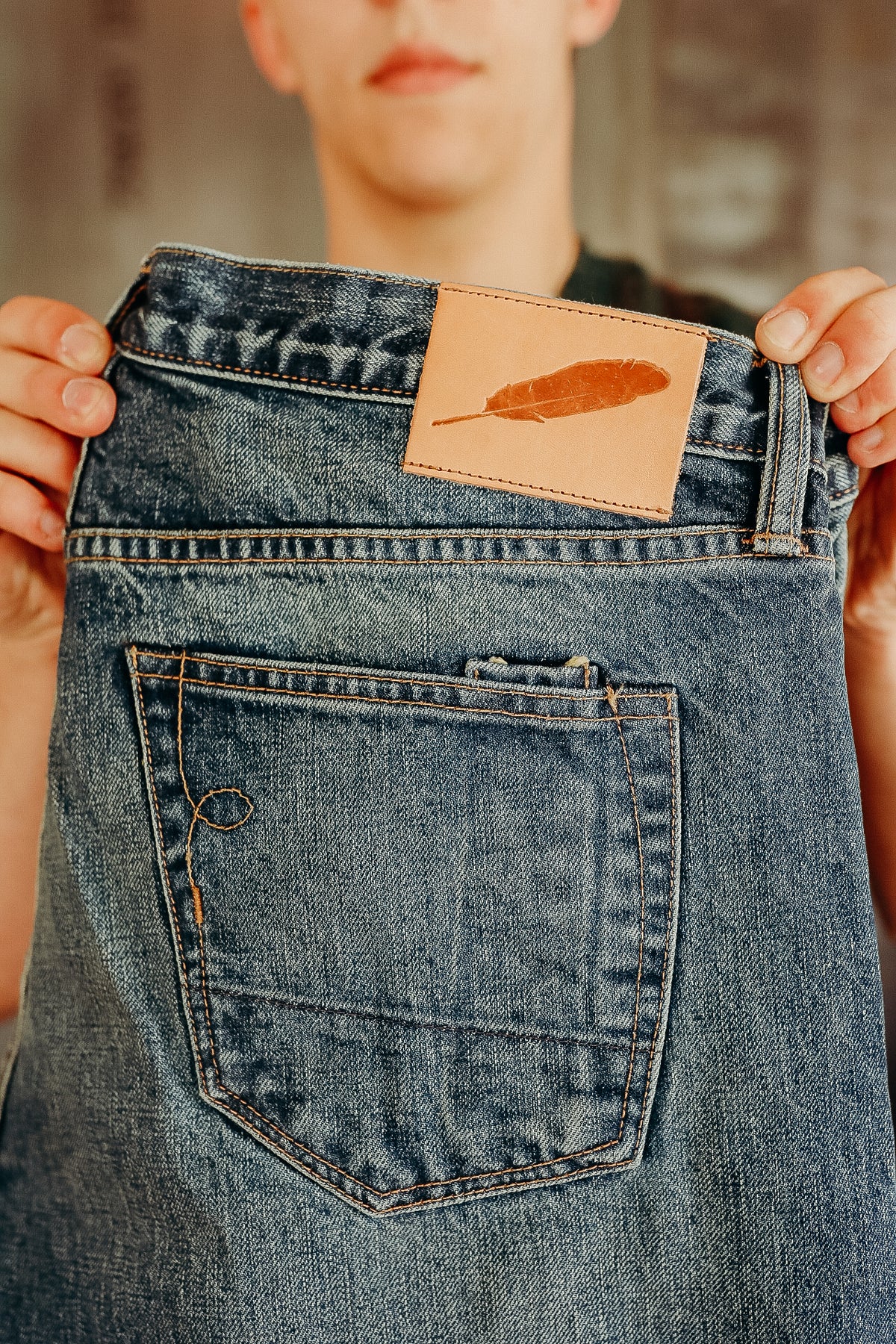 Person holding blue denim jeans with a leather patch featuring a feather logo at the waistband