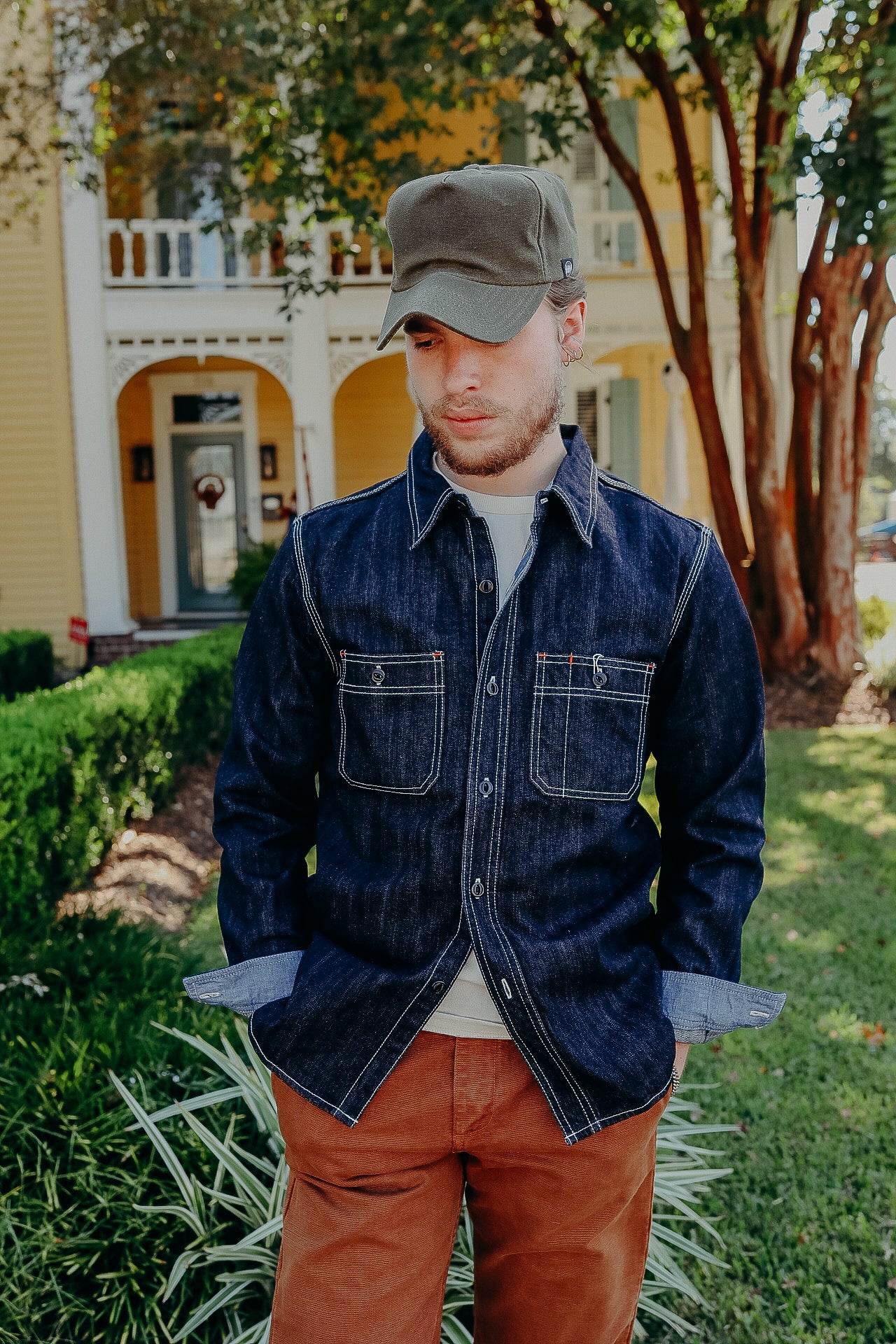 Man wearing dark denim jacket, olive green cap, and brown pants standing in front of yellow house with porch