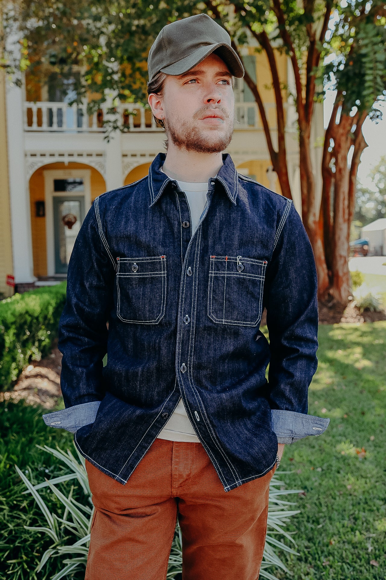 Man wearing dark denim shirt with contrast stitching, rust-colored pants, and green cap outdoors in front of yellow house