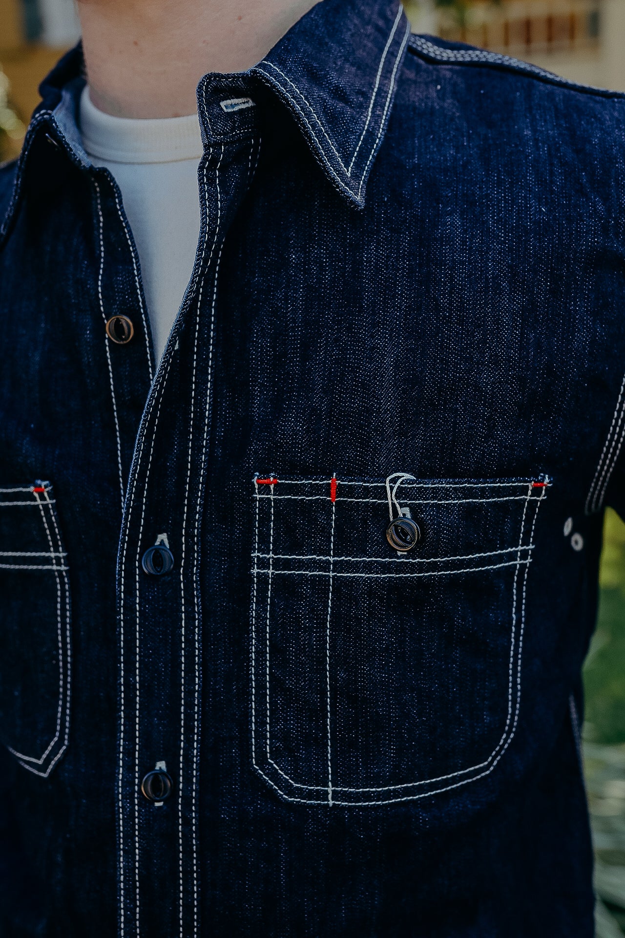 Close-up of a dark blue denim work shirt with contrast white stitching and chest pockets, worn over a white undershirt