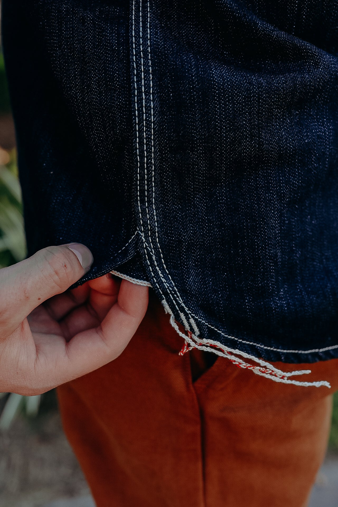 Close-up of hand holding dark blue denim jeans hem with white and red stitching detail over brown pants