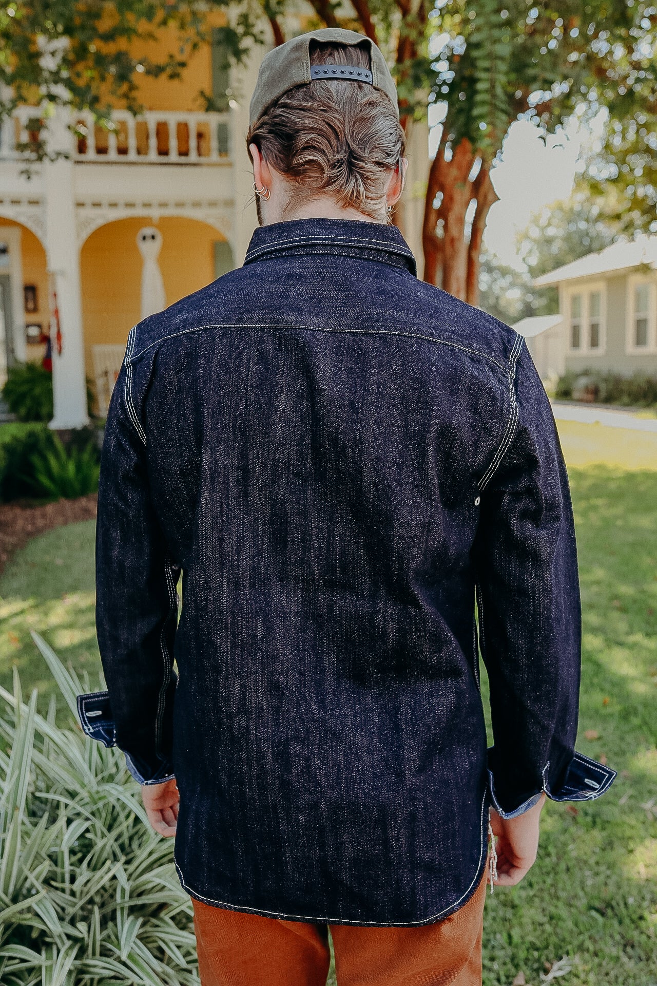 Man wearing dark indigo denim jacket and brown pants standing outdoors near house and greenery