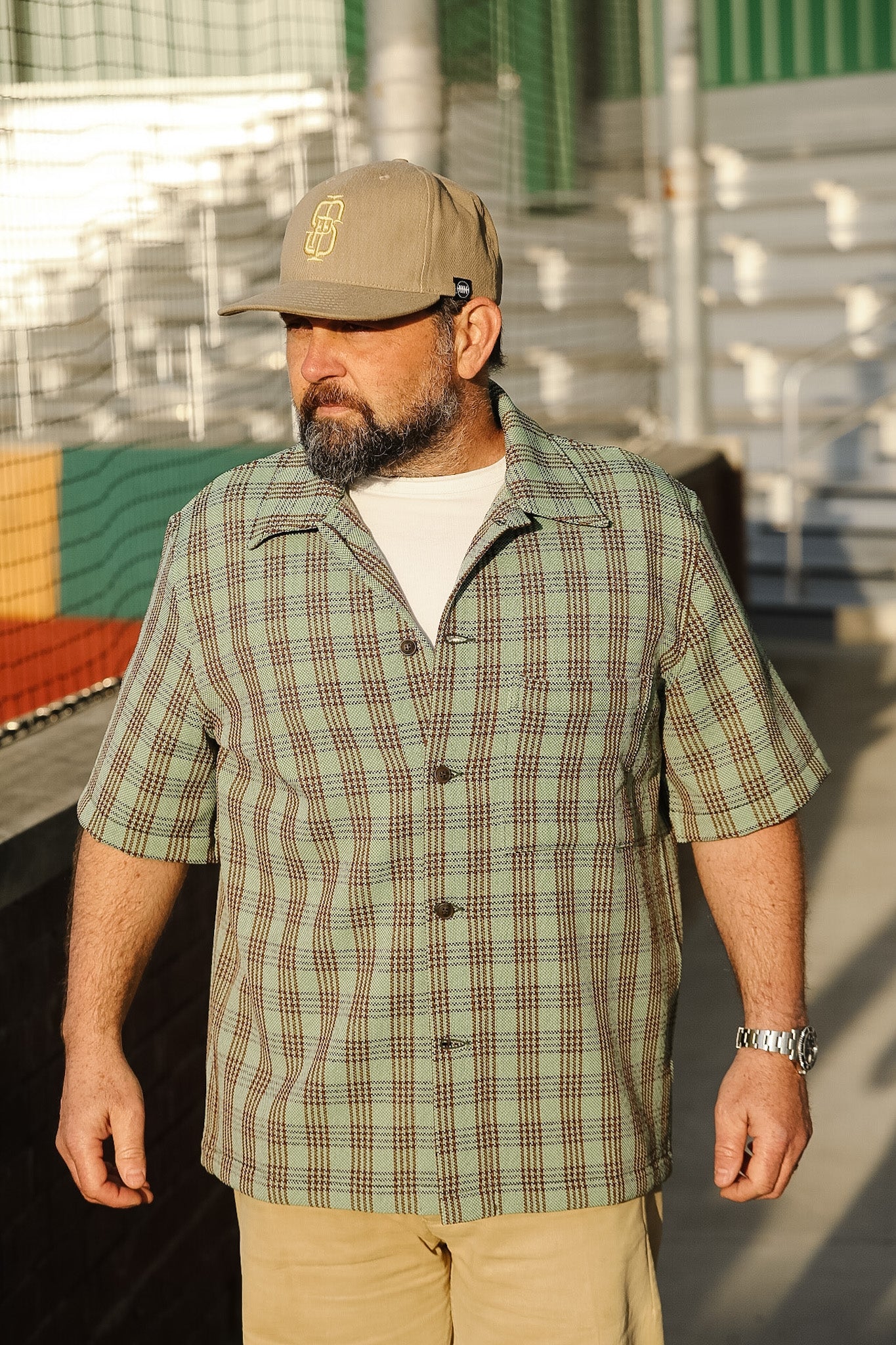 Man wearing a plaid shirt and cap standing outdoors with stadium seats in the background