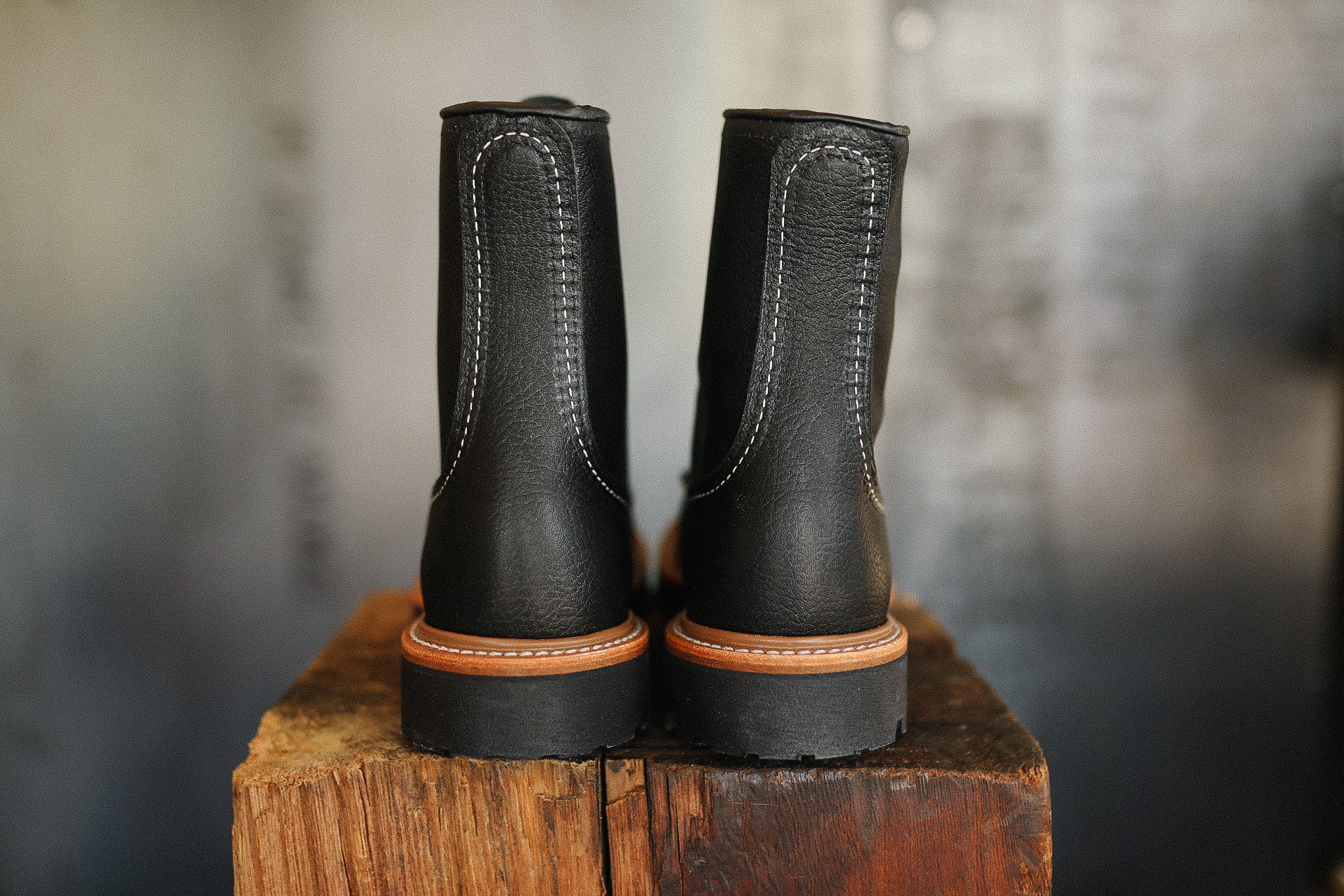 Rear view of black leather work boots with white stitching and brown trim on wooden surface